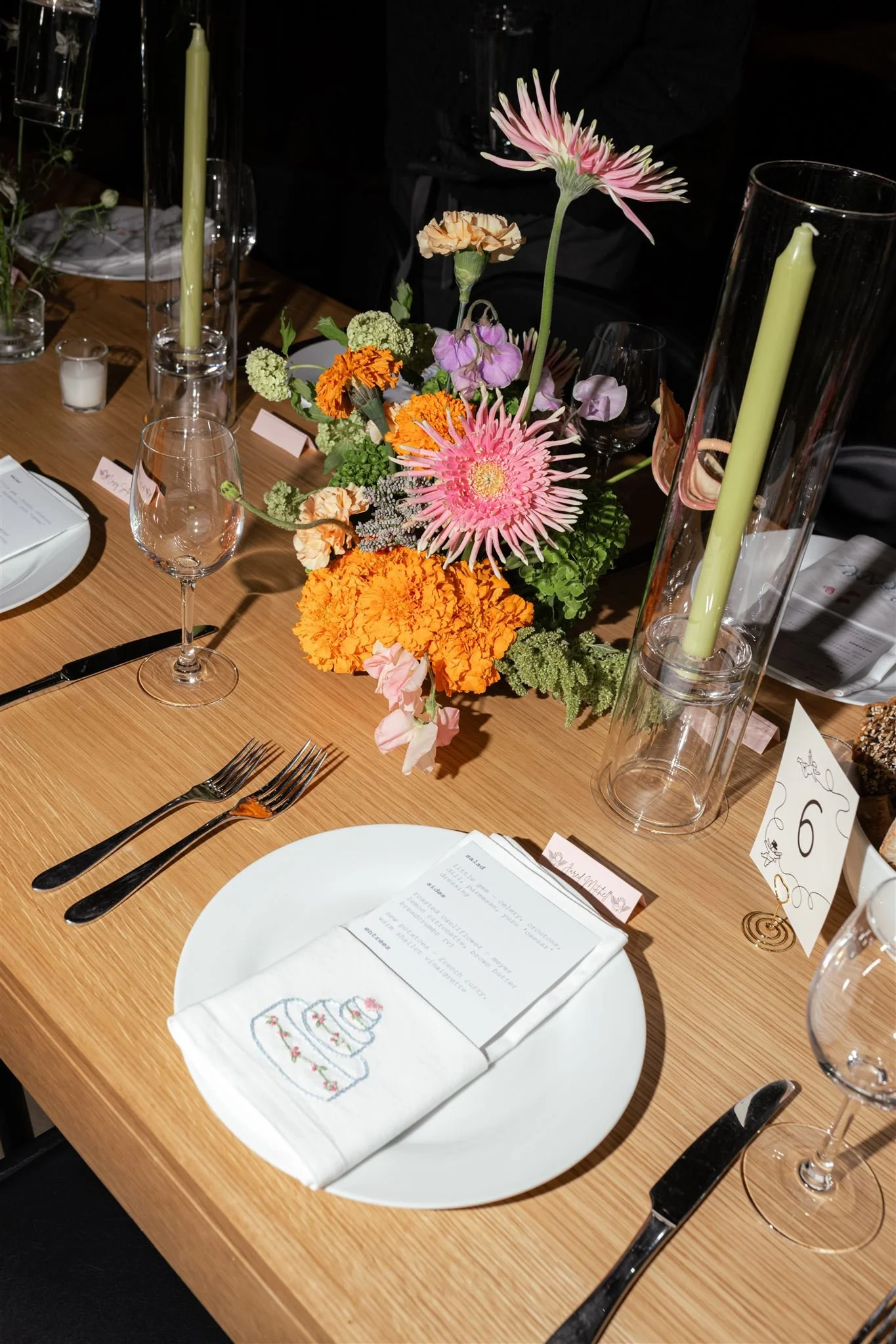 A dinner table setting with a floral centerpiece, candles, wine glasses, and a place setting with a printed menu and a napkin embroidered with a cake design.