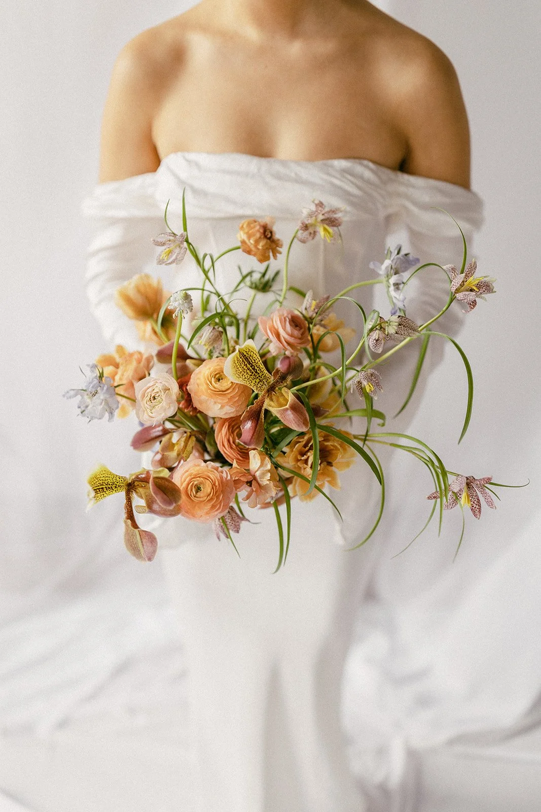 A woman in a white off-shoulder dress holding a bouquet of pastel-colored flowers including roses, orchids, and other blooms.