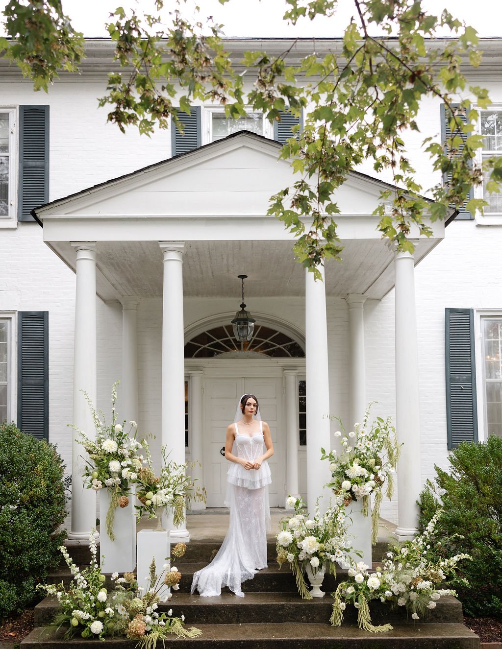 A woman in a white wedding dress standing on the steps of a white house with a portico and columns, surrounded by white floral arrangements.
