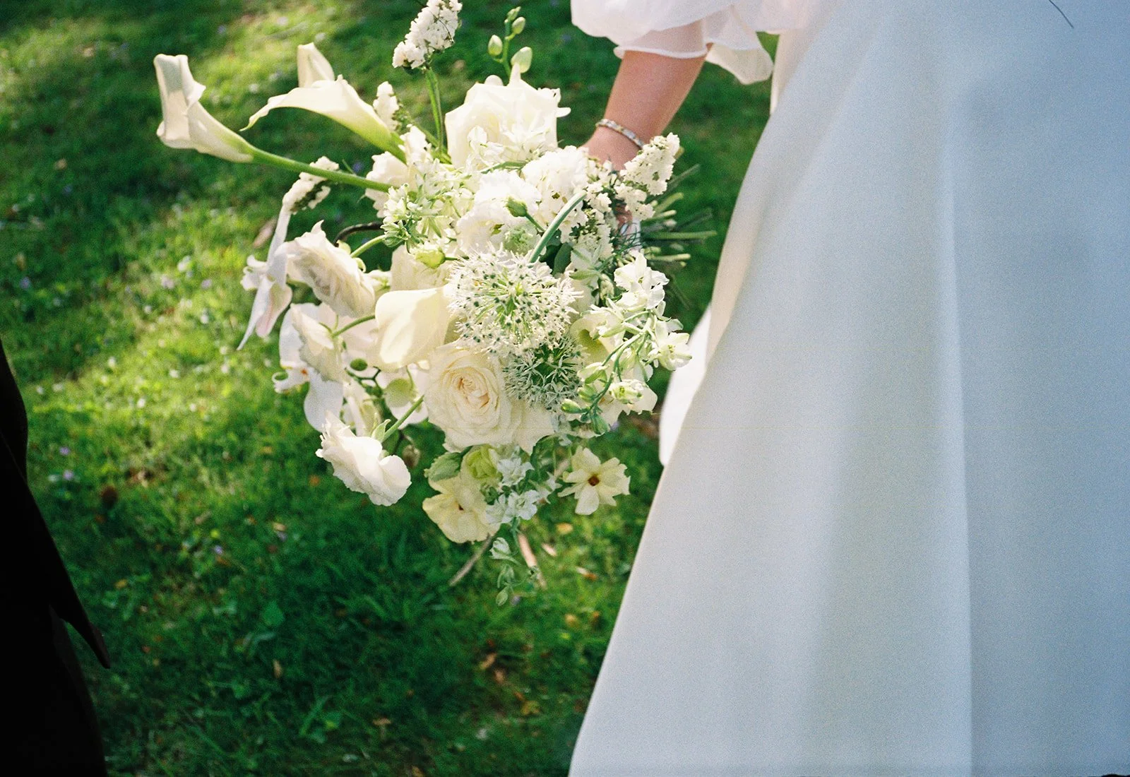 Close-up of a bride holding a bouquet of white flowers, including roses, calla lilies, and other blooms, with a blurred green grass background.