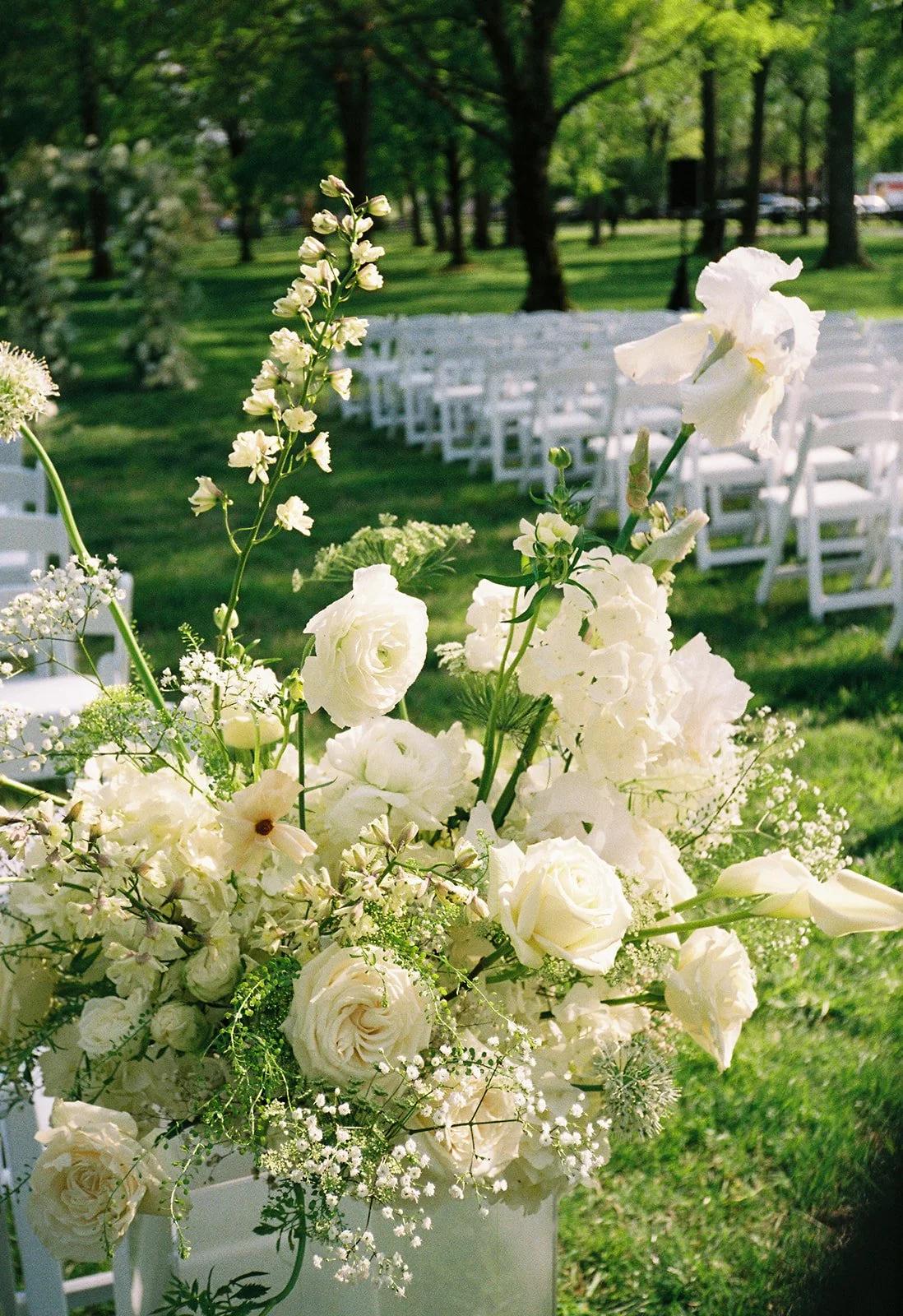 A bouquet of white flowers on an outdoor wedding ceremony setup with white chairs and green trees in the background.