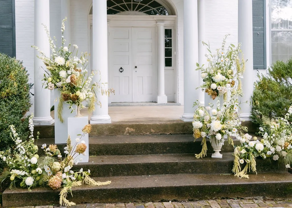 White floral arrangements on porch stairs of a white house with columns.