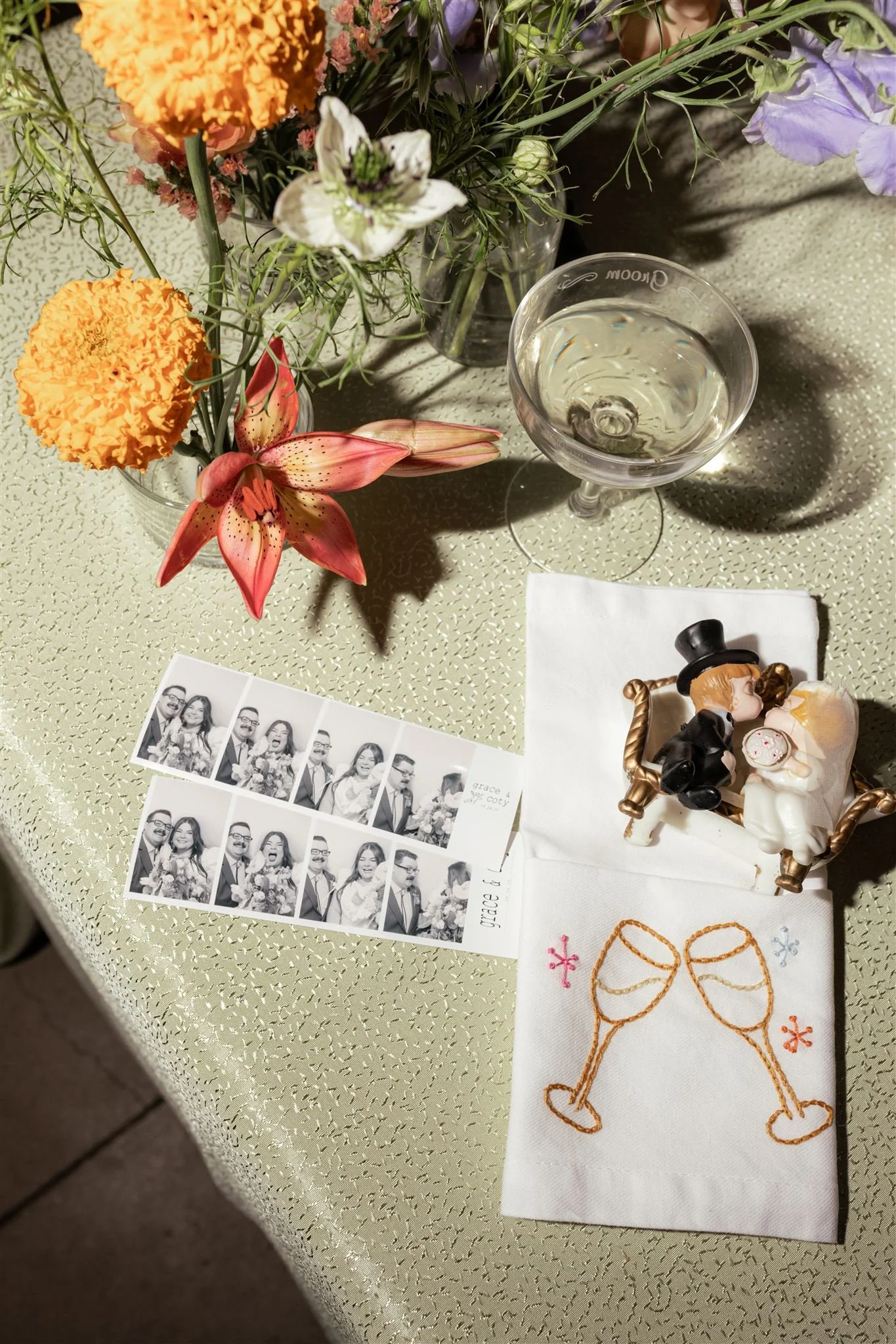 A table decorated with a bouquet of flowers, a glass of white wine, a photo strip, and a napkin with embroidered champagne glasses and colored flowers, all on a textured cream-colored tablecloth.