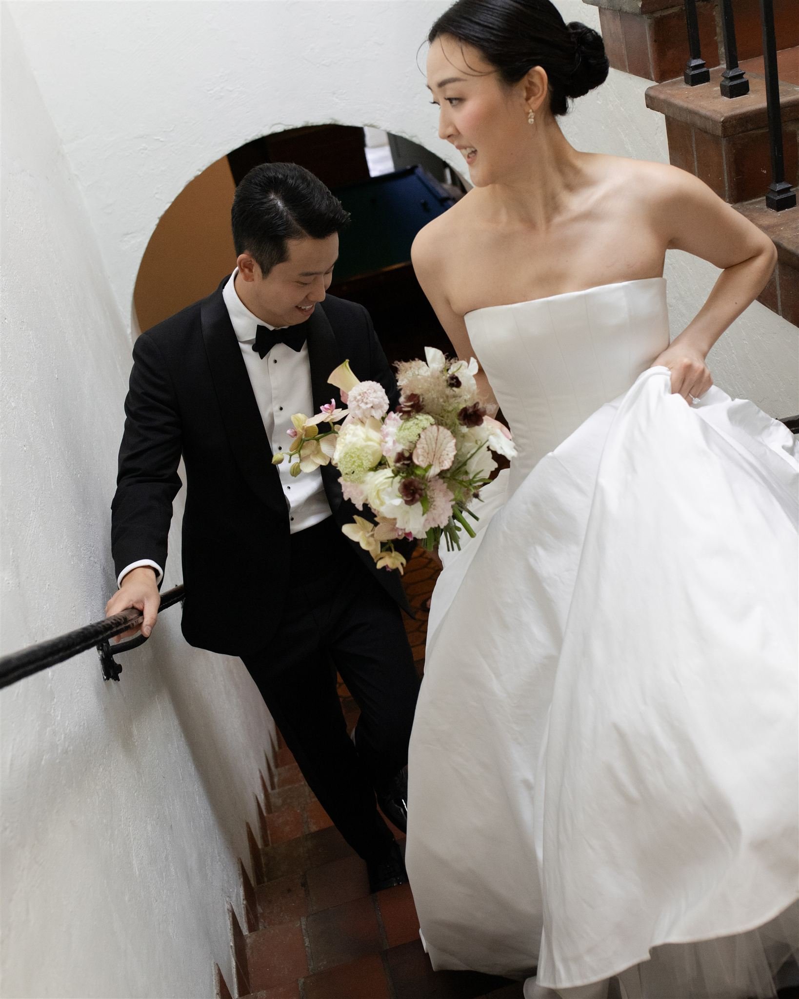 A bride in a white strapless wedding gown holding a bouquet of flowers, smiling at a groom in a black tuxedo as they walk down the stairs together.