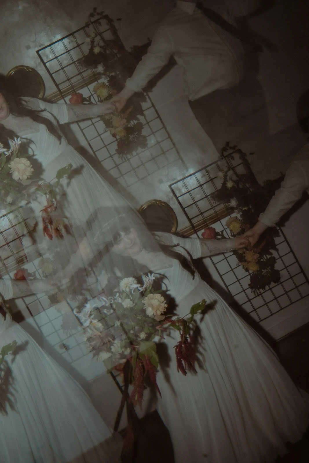 A woman in a white dress is holding hands with a man in a light-colored shirt. They are standing in front of floral and greenery decorations on metal grids, with additional floral arrangements near the woman. The photo appears to be taken indoors wit