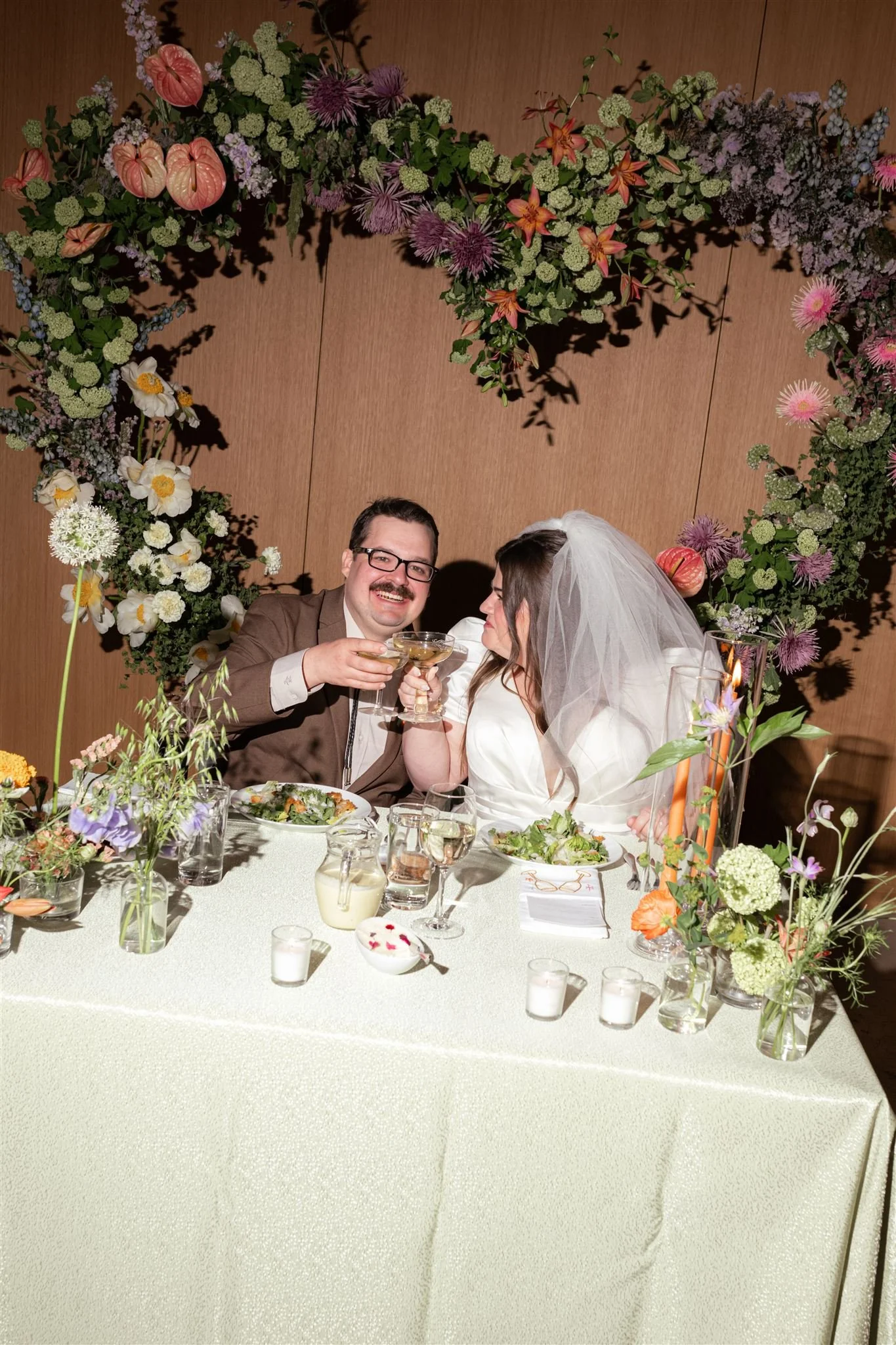 A bride and groom at a wedding reception celebrating with cocktails, sitting at a table decorated with flowers and candles, with a floral heart-shaped backdrop.