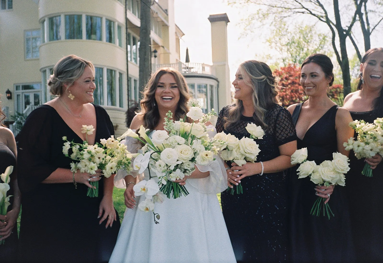 A group of women in formal dresses smiling and holding bouquets of white flowers outdoors, with a large house or mansion in the background.