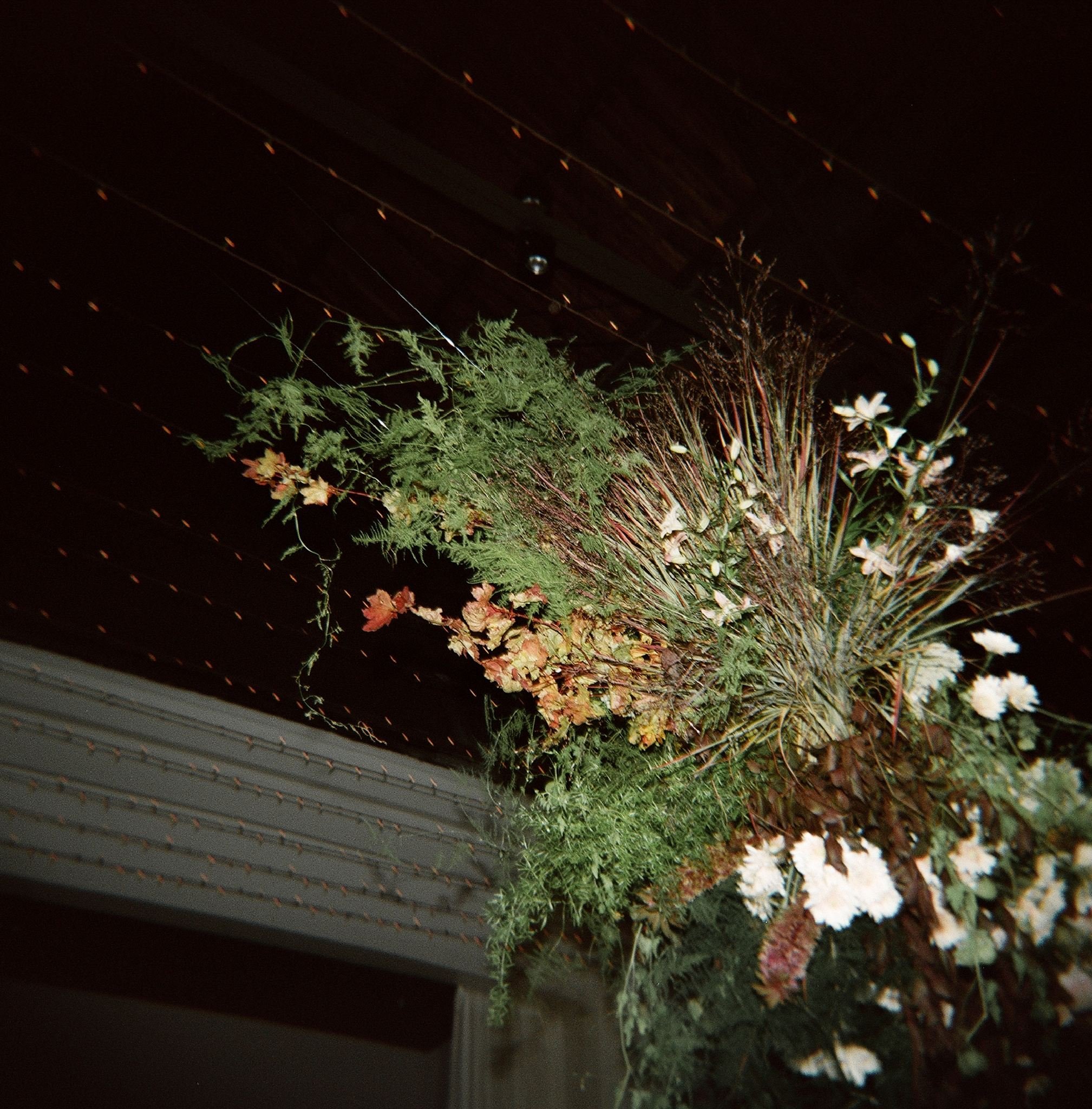 A hanging floral arrangement with white flowers and various green and brown foliage, suspended from a dark ceiling with small yellow lights.