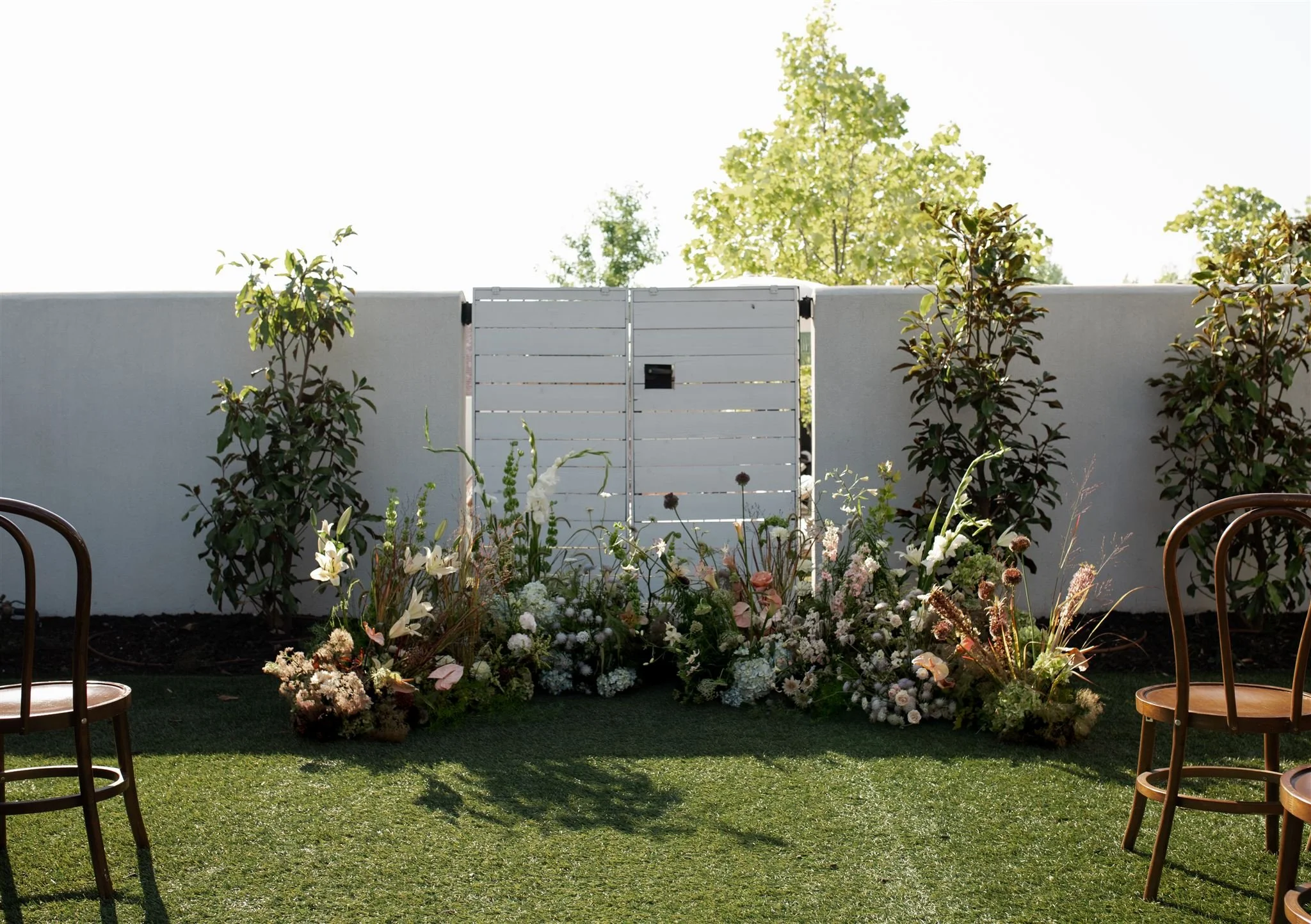 A small outdoor garden with a white wooden gate, surrounded by flower arrangements, and chairs on a green lawn, with a white wall and trees in the background.