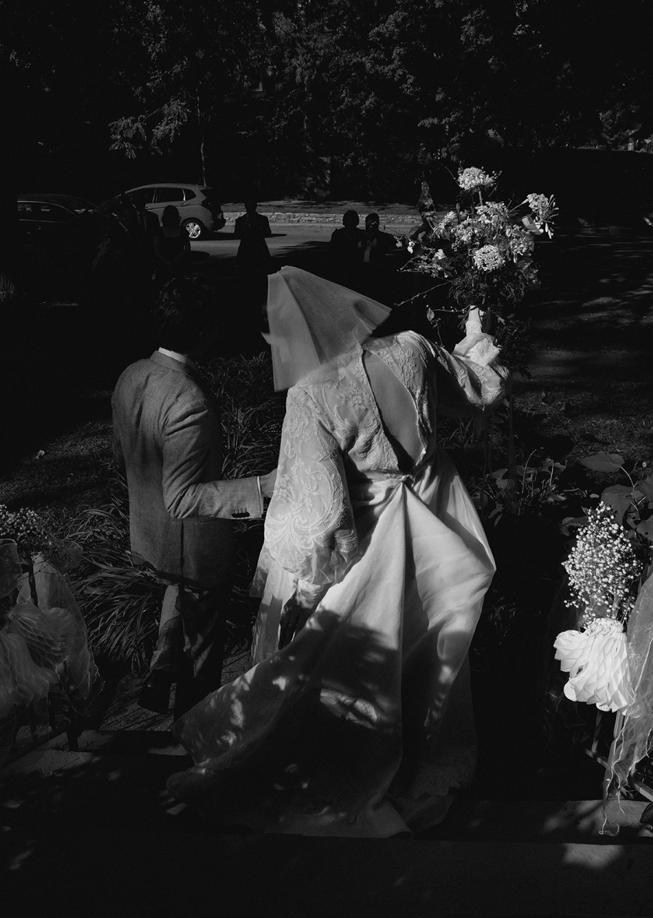 A black and white photo of a wedding ceremony at night, featuring a bride in a lace gown and a face covering, holding a bouquet, standing next to a man in a suit. Other people are visible in the background near parked cars.