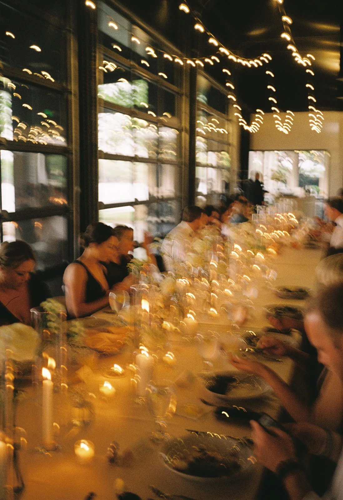 A long dinner table in a restaurant with multiple guests eating, with string lights hanging from the ceiling and candles on the table, creating a cozy atmosphere.
