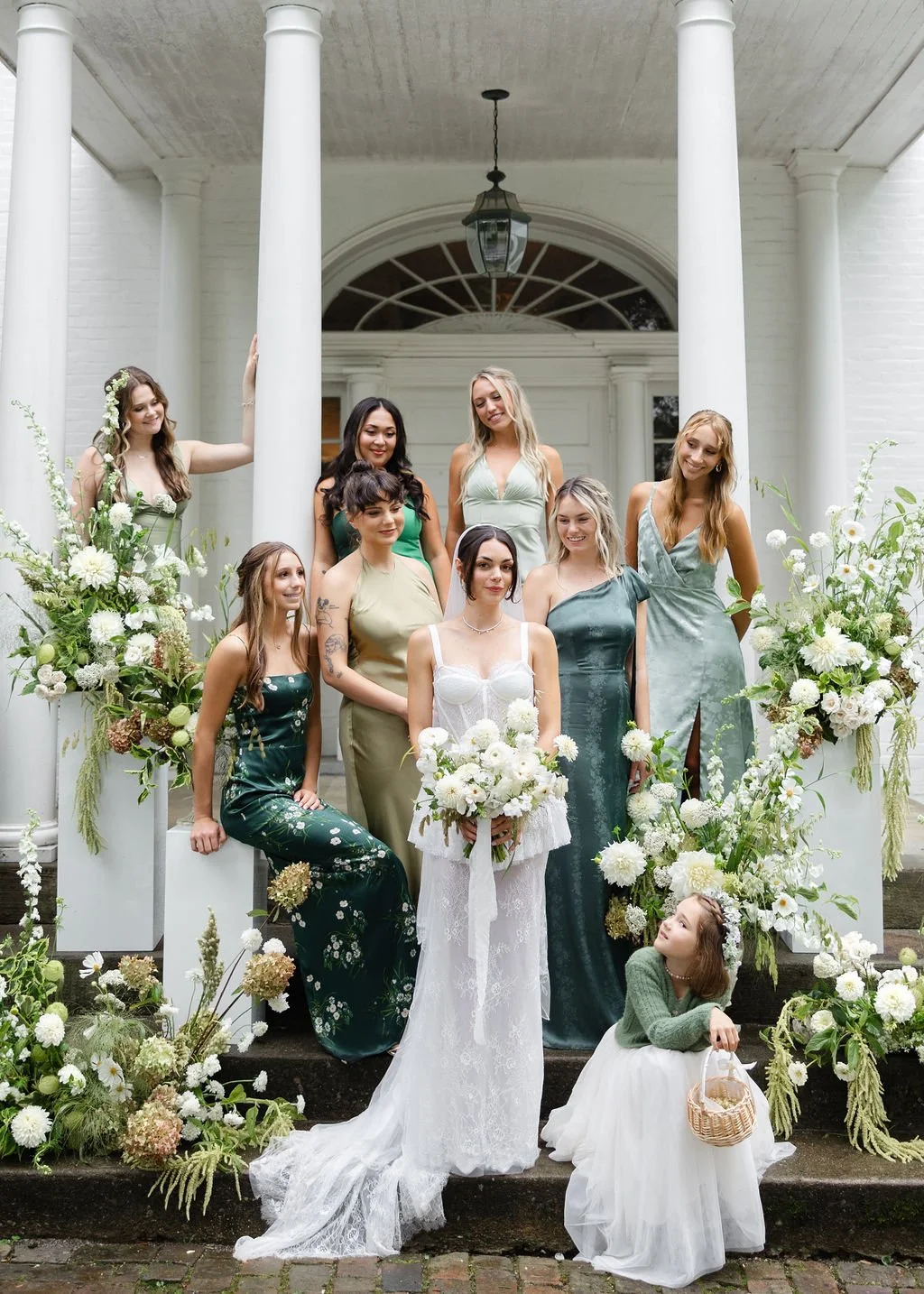 A bridal party of nine women, including a bride in a white dress holding a bouquet, and a young girl in a green sweater and white tulle skirt with a basket, standing on or in front of the steps of a white house with columns, surrounded by white and g