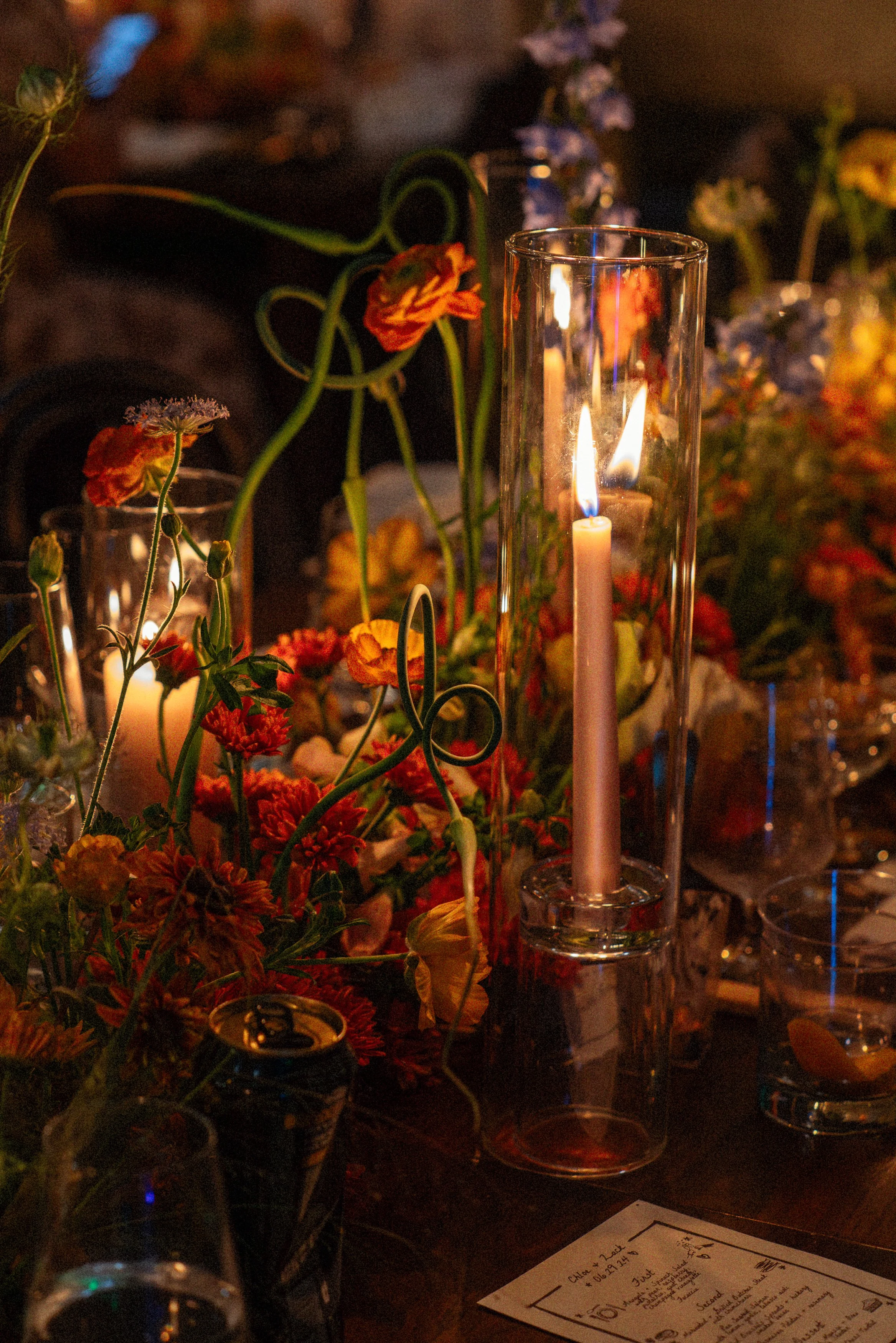 Tall glass candleholder with lit pink taper candle on a decorated table with flowers and drinks.