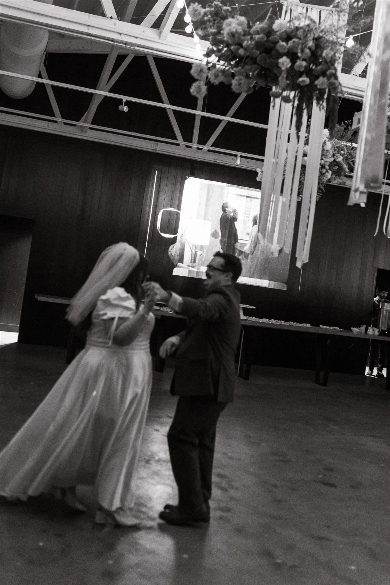 A bride and groom dancing at a wedding reception, with the bride wearing a white gown and veil and the groom in a suit. They are holding hands and smiling. The background features a dark wall, a large floral arrangement hanging from the ceiling, and 