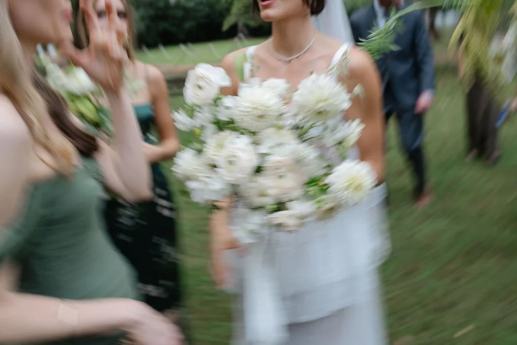 Blurry image of a woman in white holding a large bouquet of white flowers at an outdoor event, surrounded by other women and people in the background.