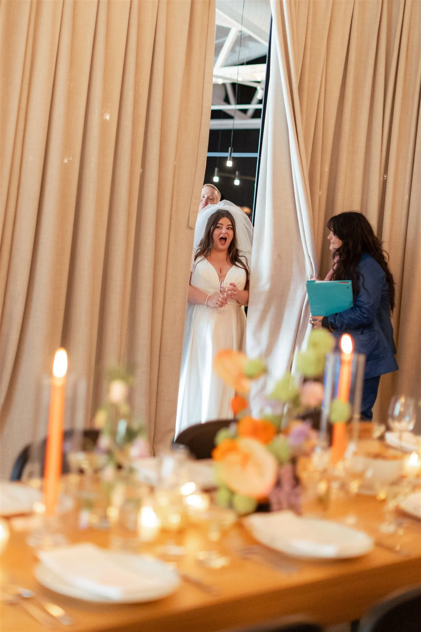 A bride in a white wedding dress and veil holding a glass, surprised as she peeks through beige curtains at a wedding reception; a woman holding folders stands nearby, smiling, and a man is partially visible behind the curtains; decorated table with 