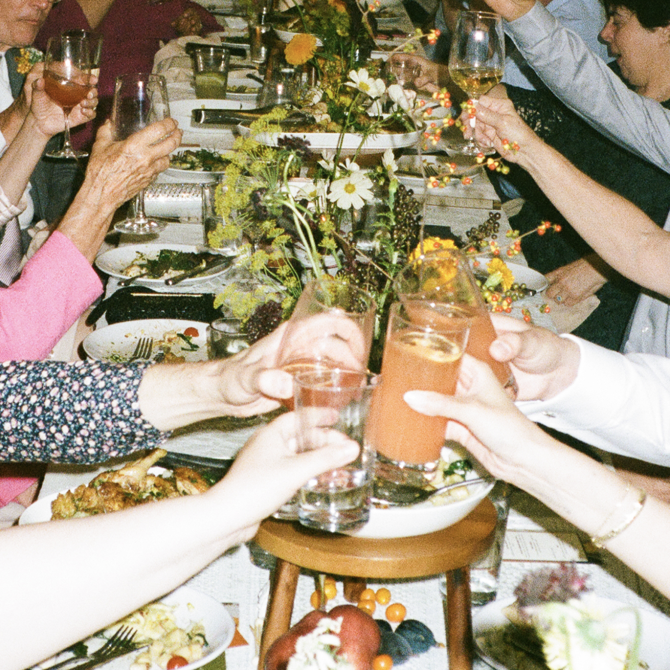 People raising glasses with drinks at a festive dinner table decorated with flowers and food.