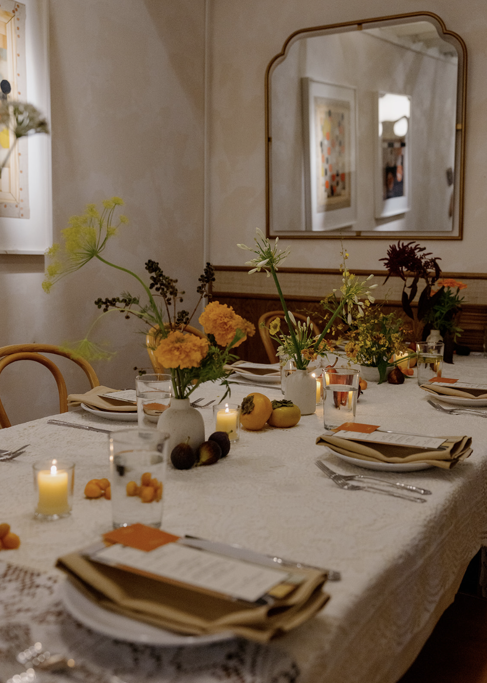 A dining table decorated with vases of flowers, candles, and fruits, set for a meal with napkins, plates, and cutlery in a cozy room with a mirror on the wall.