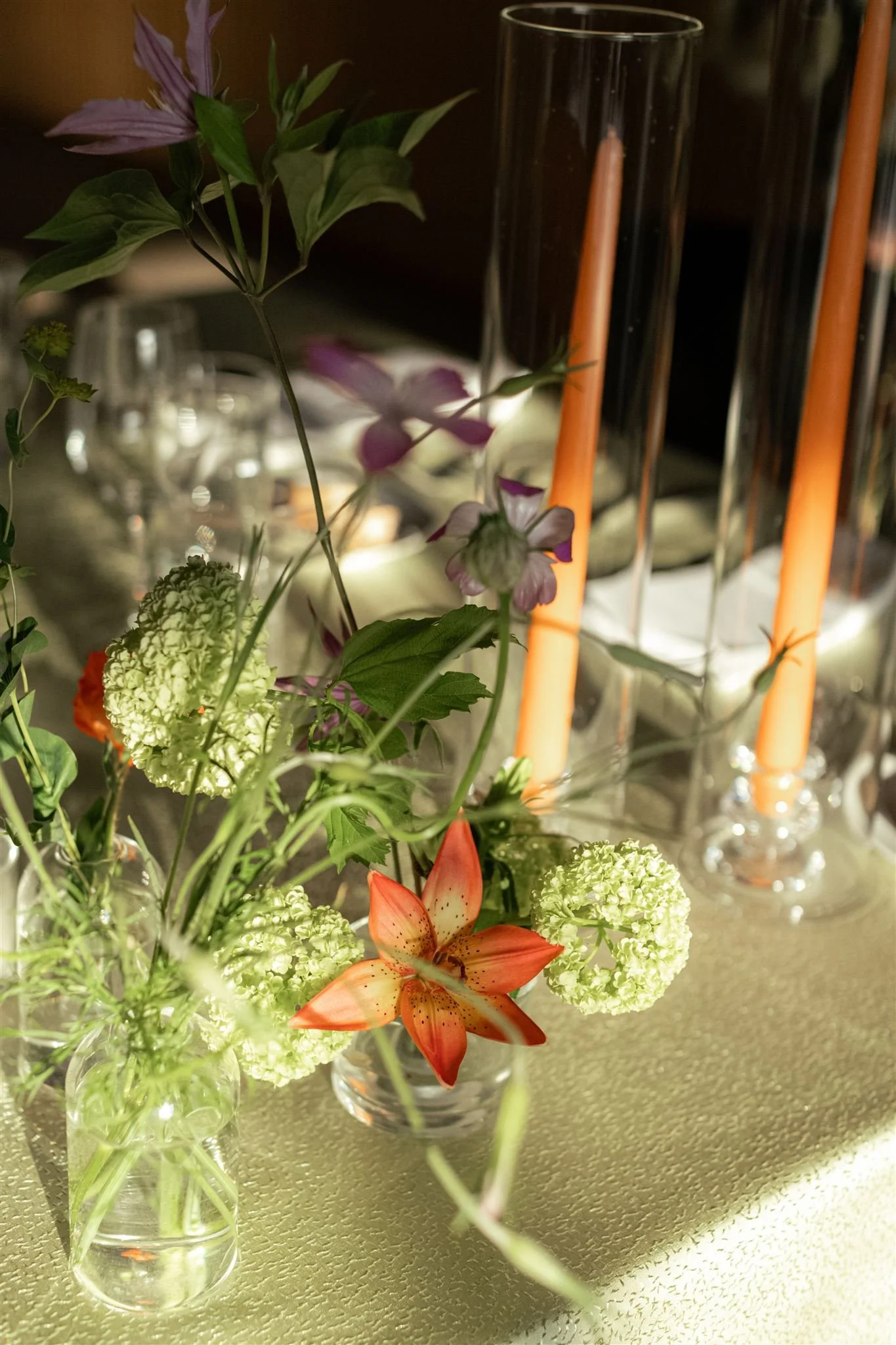 A floral centerpiece with a variety of flowers including lilies, hydrangeas, and other blooms in small glass vases on a table with tall orange candles in glass holders.