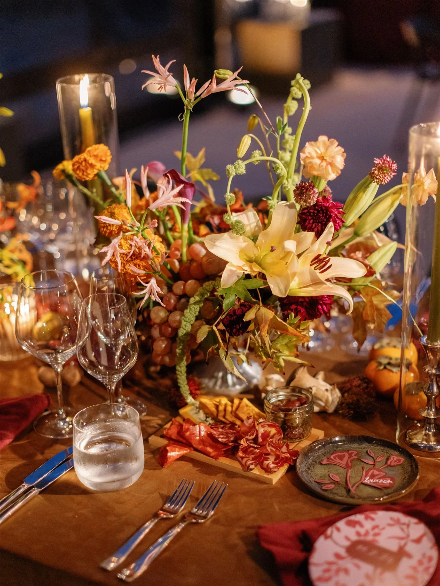 A table decorated with a tall floral centerpiece of flowers and grapes, surrounded by wine glasses, a glass of water, charcuterie, and candles.