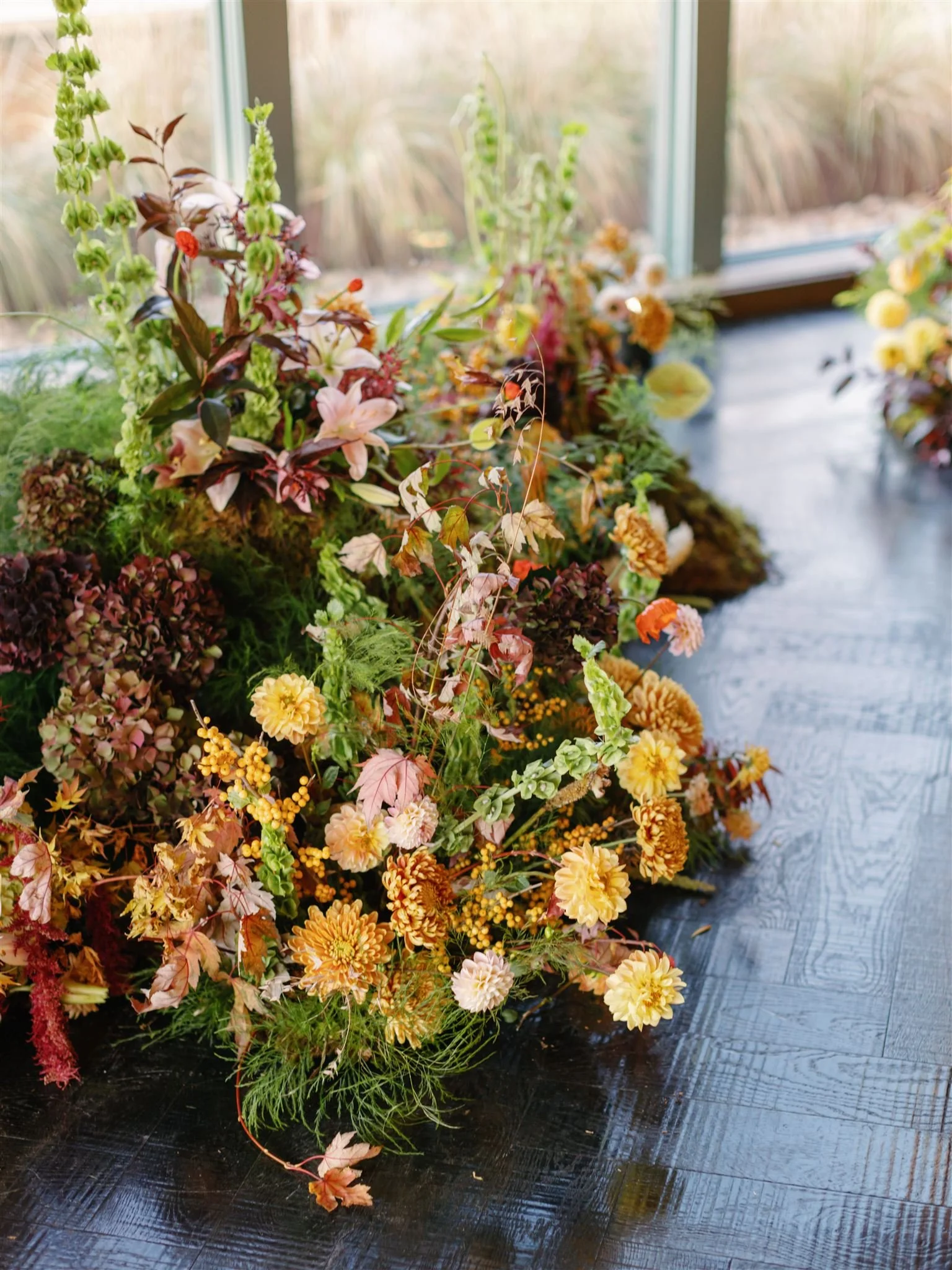 An arrangement of various flowers and greenery on a dark wooden surface near a window, with a blurred garden view outside.