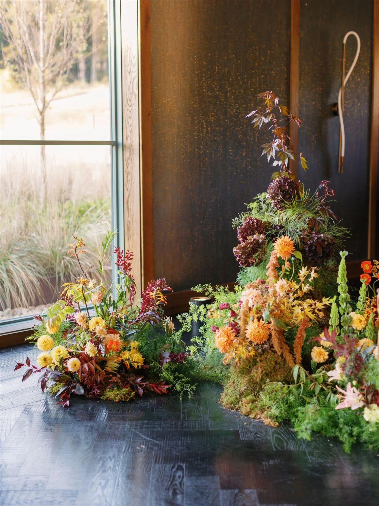 Arranged floral display of various colorful flowers and greenery on a dark wooden table near a window with outdoor view.