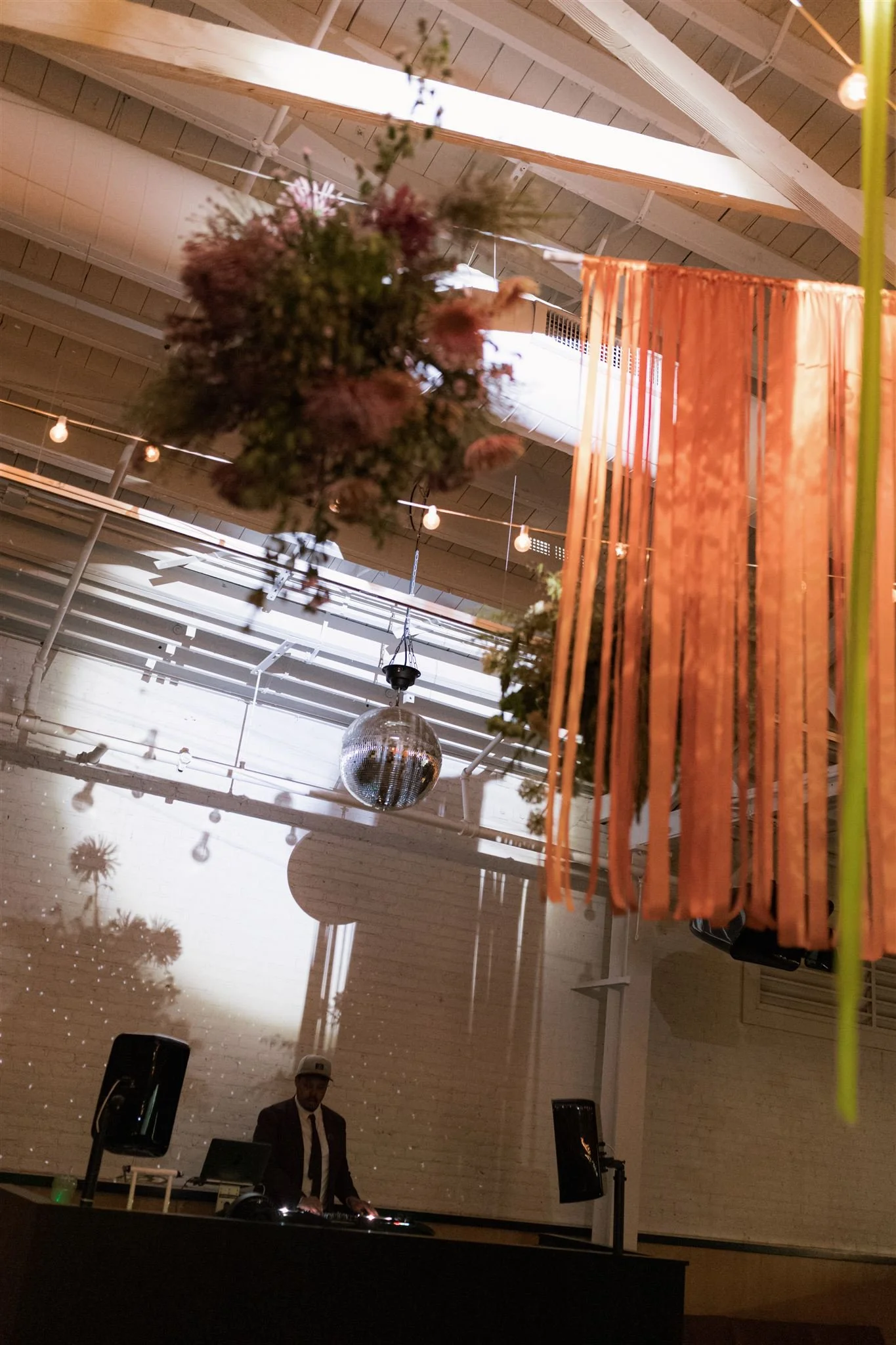 A DJ performing at a venue with hanging floral arrangements, a disco ball, and colorful hanging ribbons.