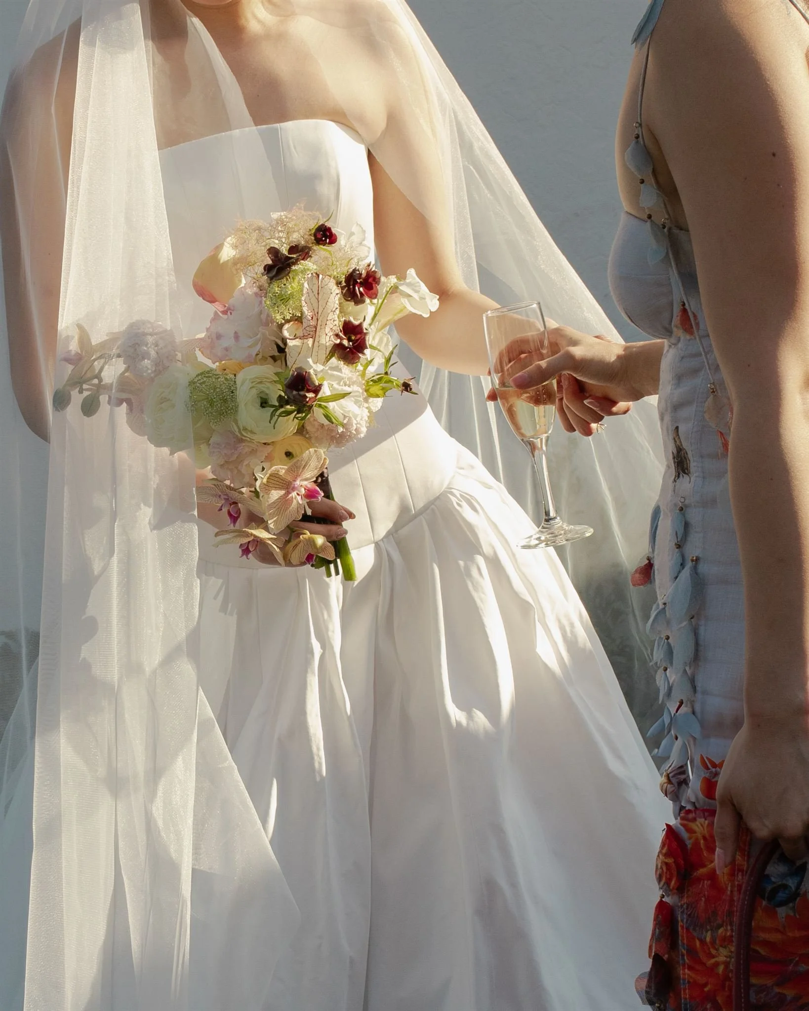 A bride in a white wedding dress holding a large bouquet of flowers, standing next to another woman holding a glass of champagne.