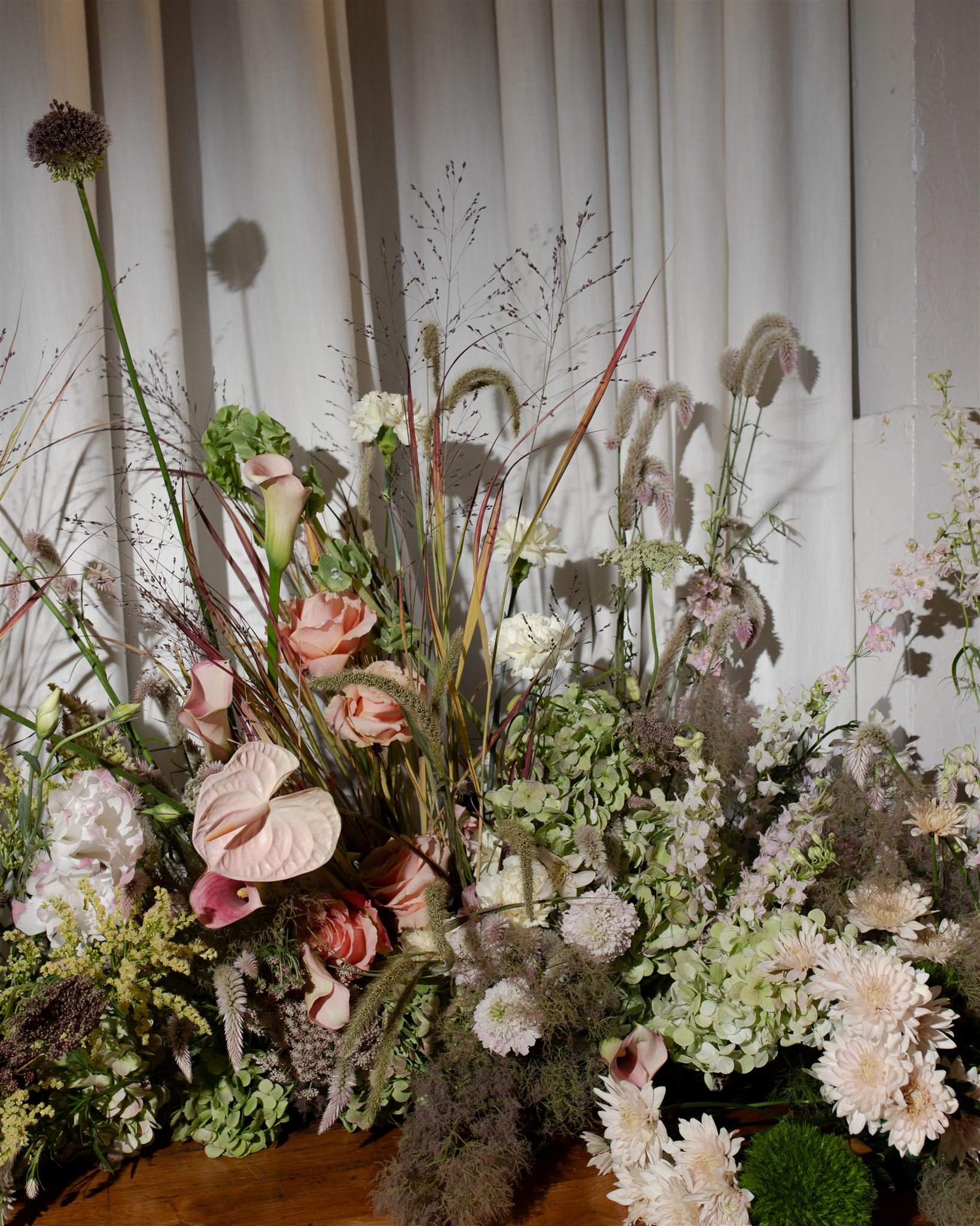 A large floral arrangement with various pink, white, and green flowers and grasses on a wooden surface against white curtains.