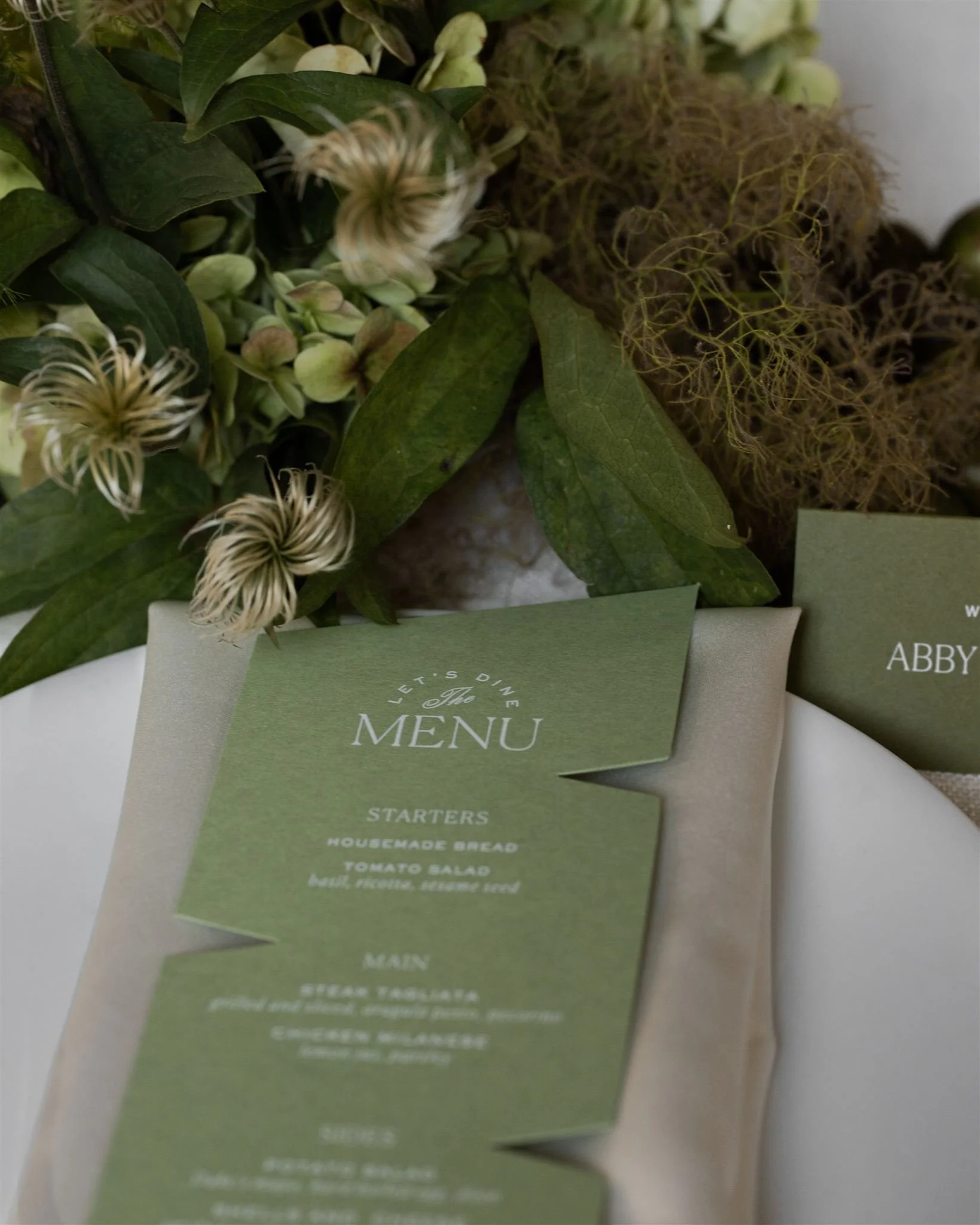 A green menu card with white text, placed on a white napkin, surrounded by green leaves, white flowers, and dried plant materials.