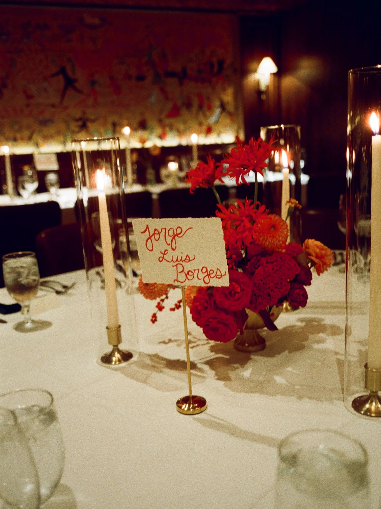 Table decorated with a floral arrangement of red and orange flowers, surrounded by tall glasses candle holders with lit candles, and a handwritten place card that reads 'Jorge & Luis Borges'.