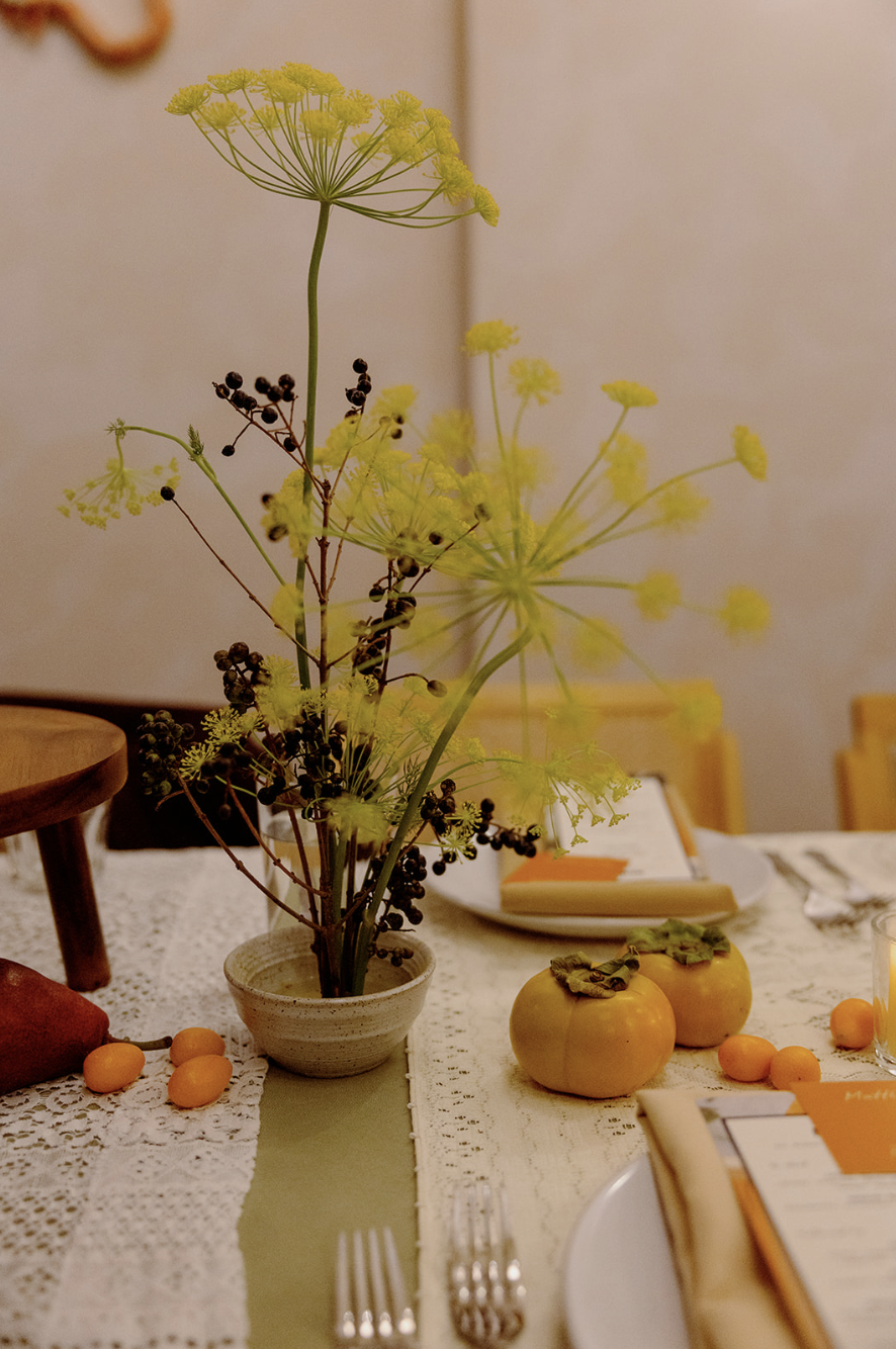 A table decorated with a vase of yellow and dark berries, two yellow decorative gourds with green leaves, and scattered yellow apricots, set for a meal with utensils, plates, and napkins.