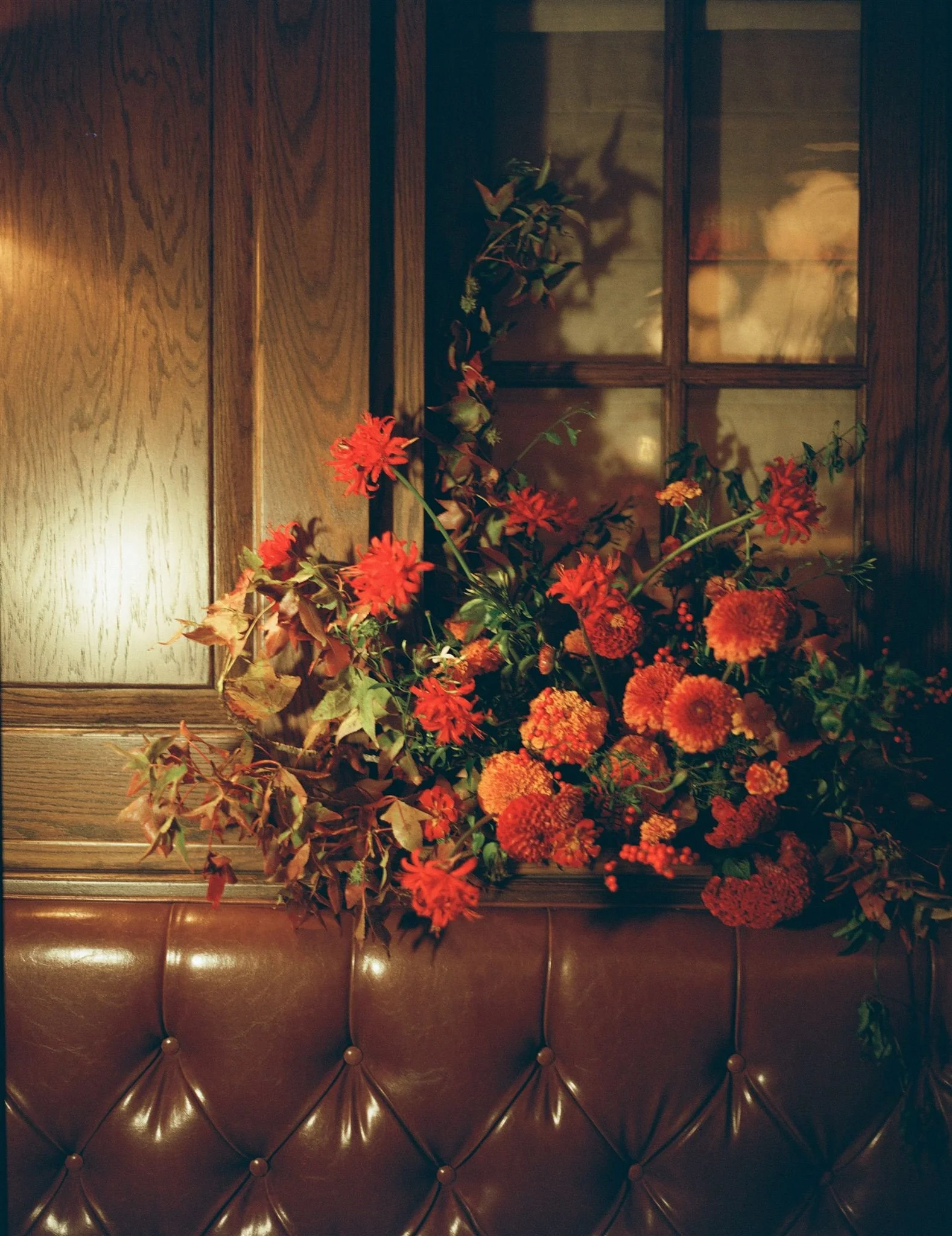 A large bouquet of red and orange flowers placed on a leather cushion against a wooden wall with a window.