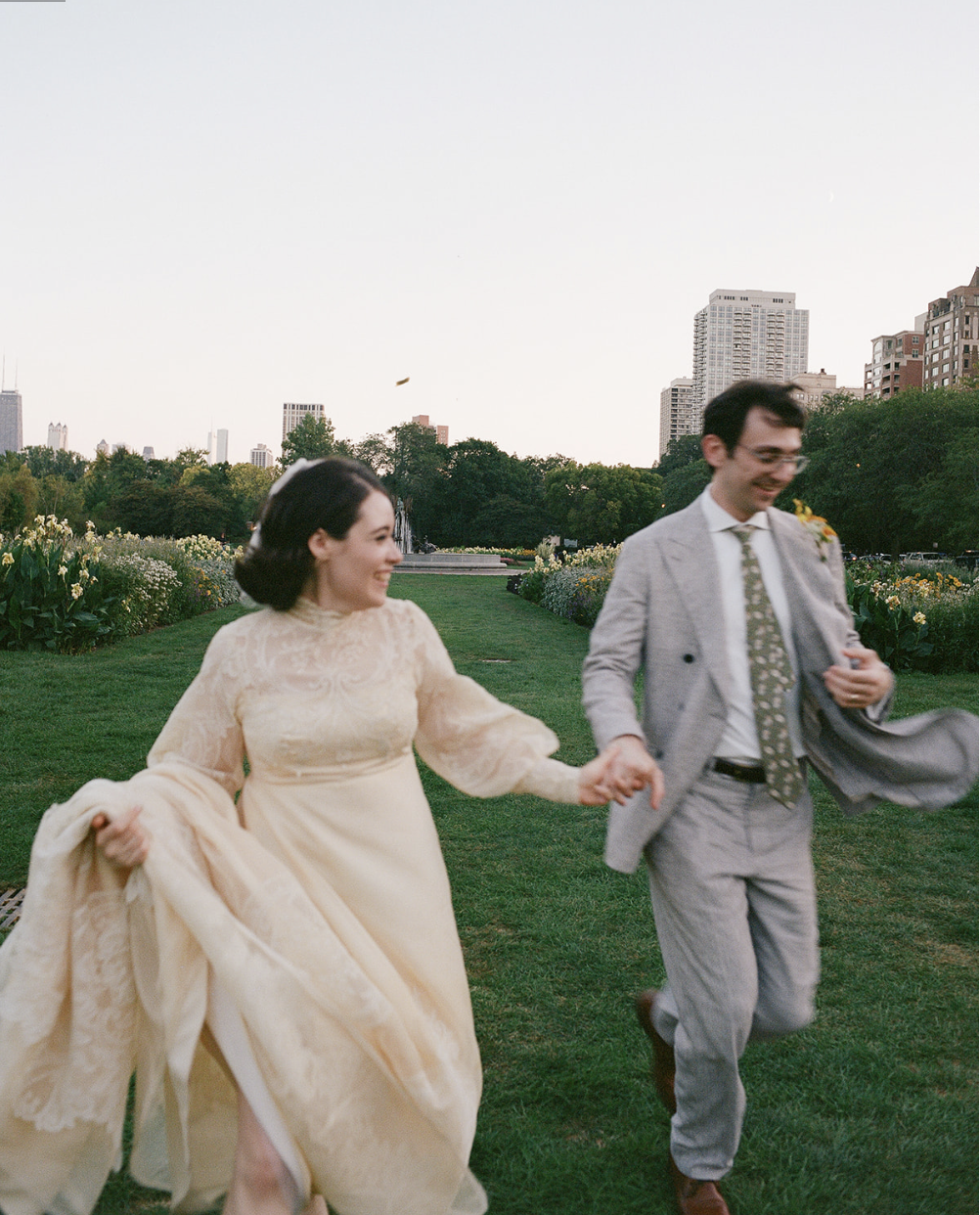 A couple holding hands and running on a grassy park with city buildings in the background during daytime.