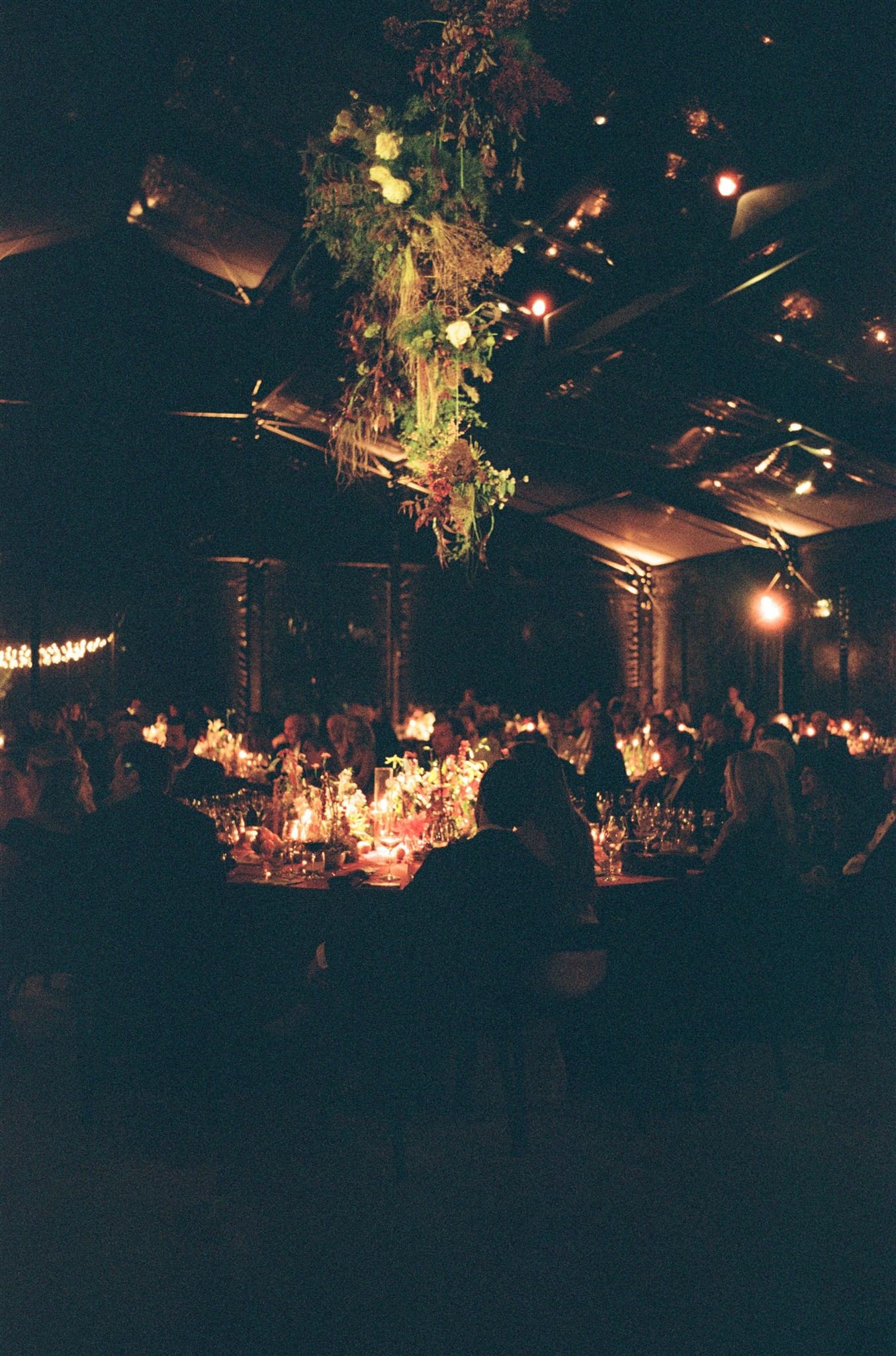 People gathered at a dimly lit banquet or wedding reception with floral centerpieces and hanging greenery arrangement in a large indoor venue.
