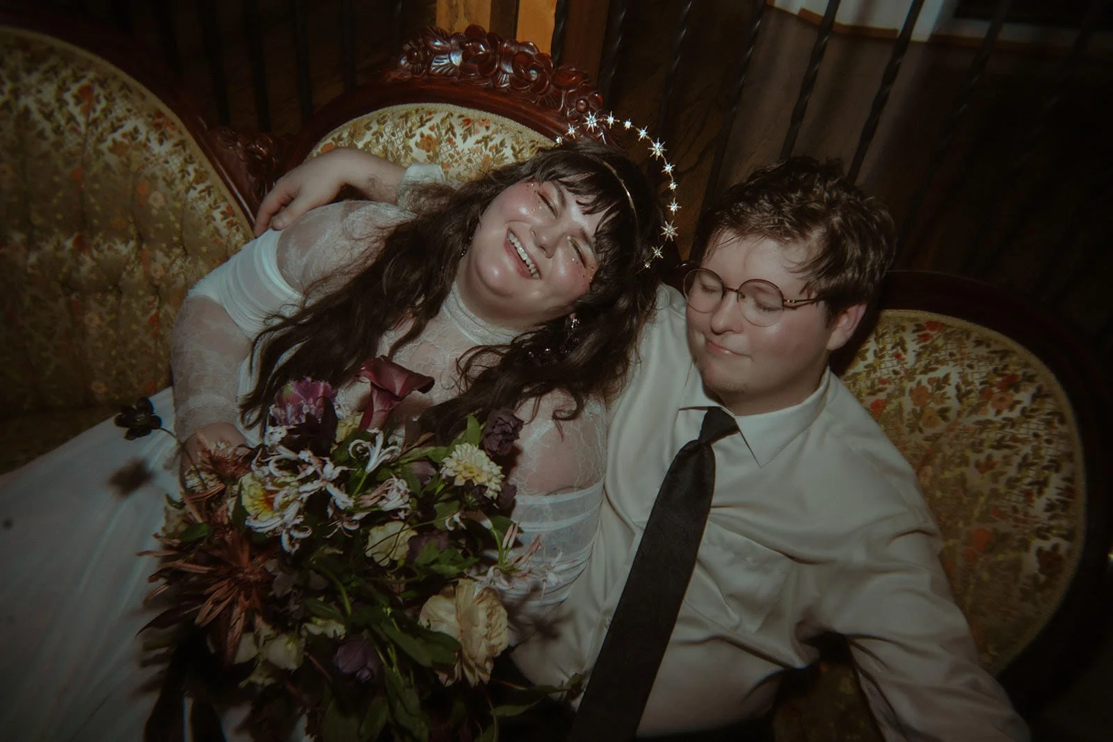 A bride and groom sitting close together on a vintage sofa, smiling and appearing happy after a wedding, with the bride holding a bouquet of flowers.