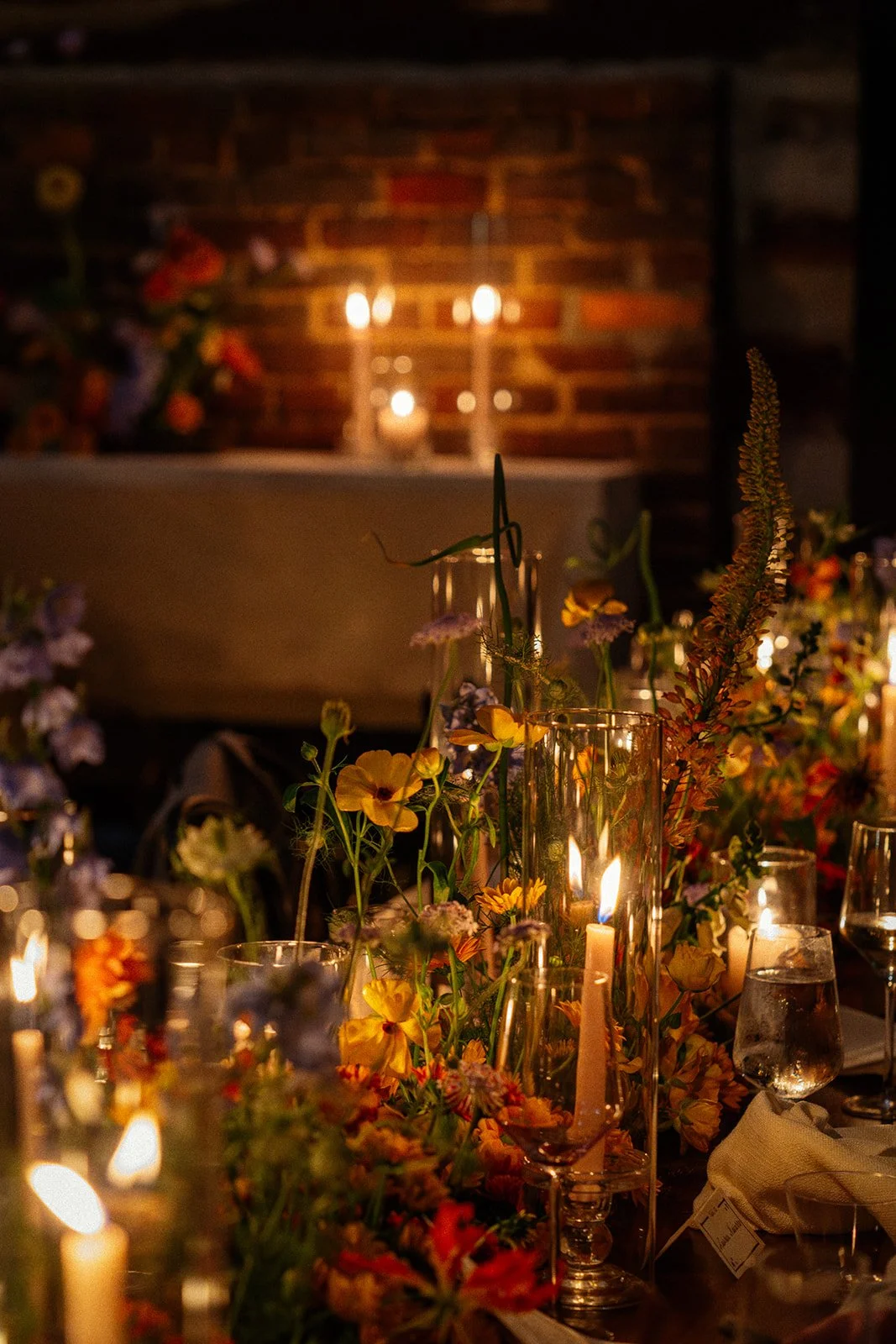 Elegant table setting with flowers and lit candles in glass holders, dimly lit with a brick wall and candlelight in the background.
