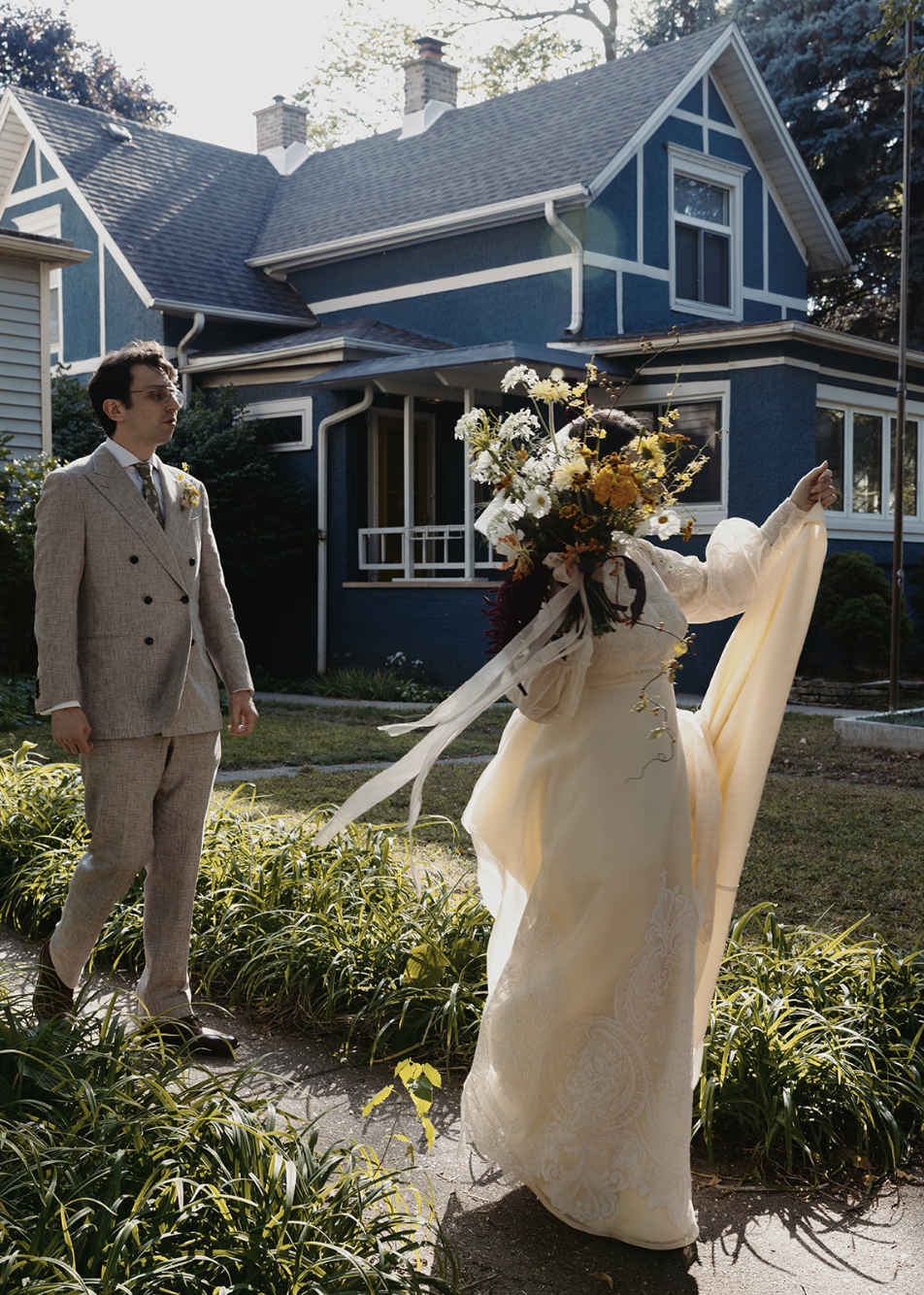 A bride holding a large bouquet of flowers and a large piece of fabric stands outside a blue house with white trim, while a man in a light-colored suit watches her.