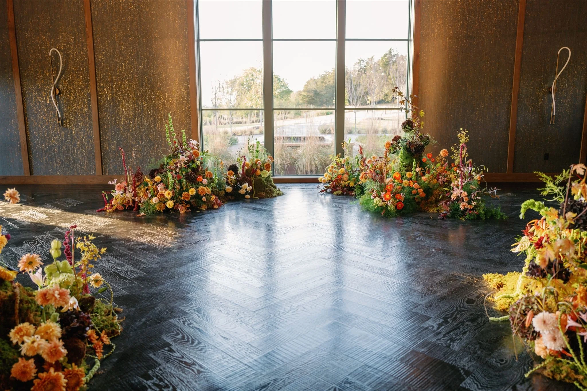An indoor space with large windows overlooking a landscape, decorated with colorful floral arrangements on a dark wooden floor.