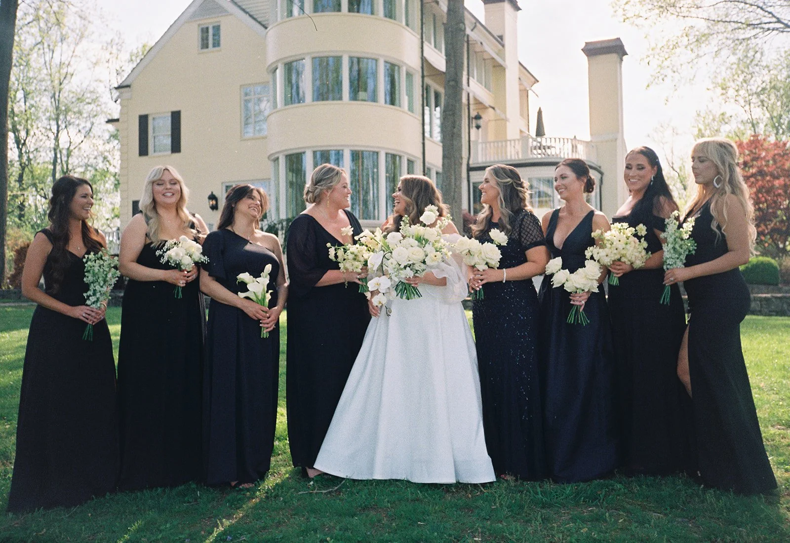 A bride and her bridesmaids standing outside on grass, holding bouquets of white flowers, in front of a large house with a rounded glass sunroom, trees, and cloudy sky.