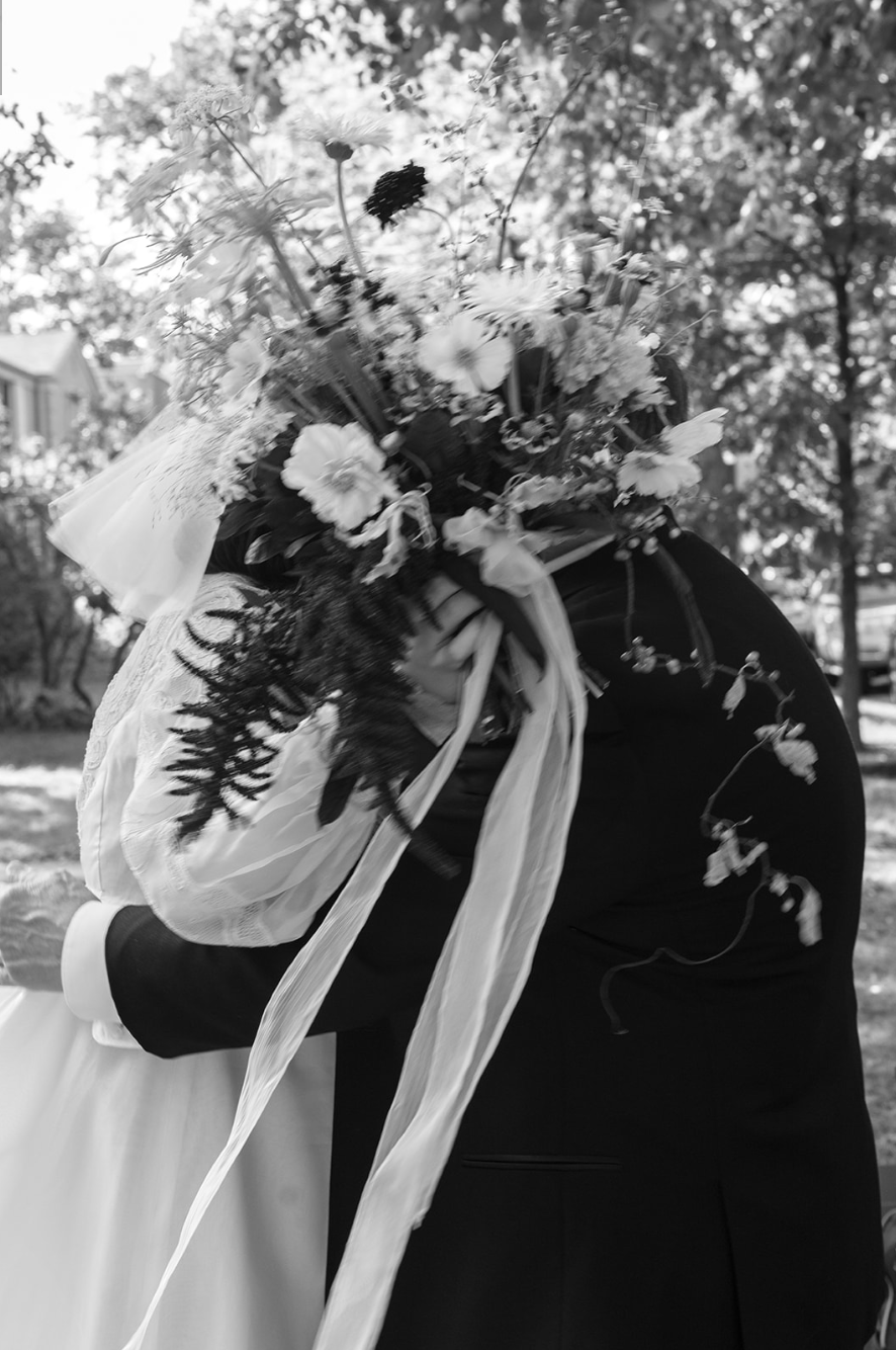 A person holding a bouquet of flowers during a wedding ceremony, outdoors with trees in the background, black and white photo.