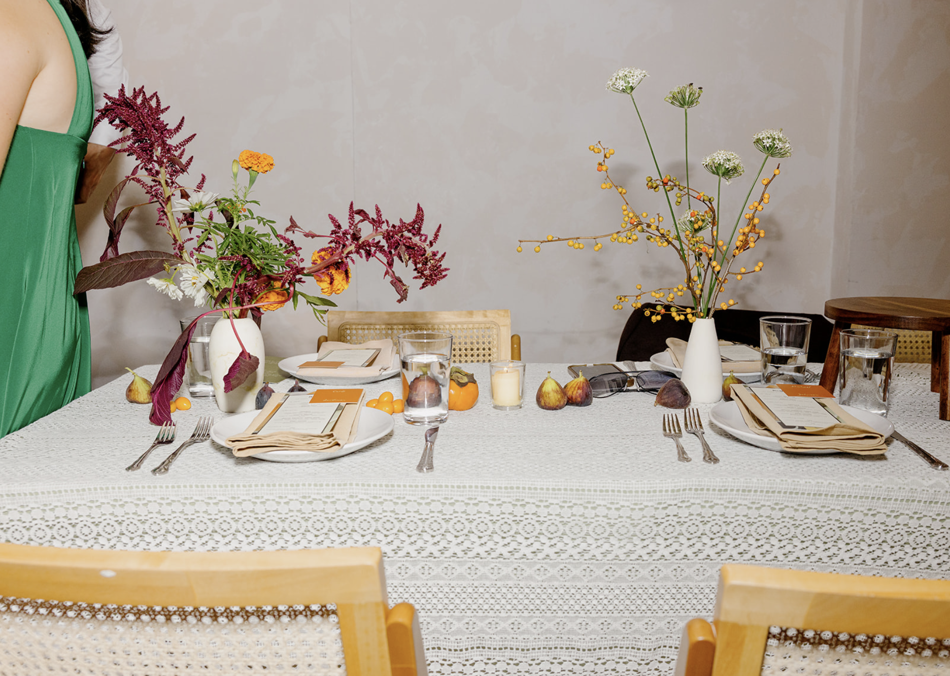 A dining table set for a meal with three large vases of colorful flowers, including purple, yellow, white, and green, and some fruits and candles, with chairs around it.
