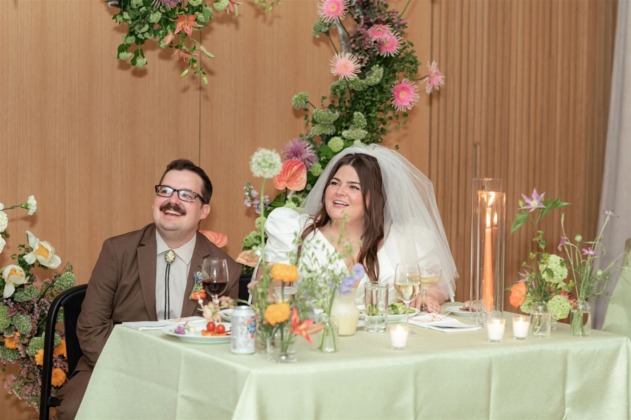 A bride and groom sit at a wedding reception table, smiling and enjoying the moment. The bride is wearing a white wedding dress and veil, while the groom is in a brown suit with glasses and a bolo tie. The table is decorated with flowers, candles, an