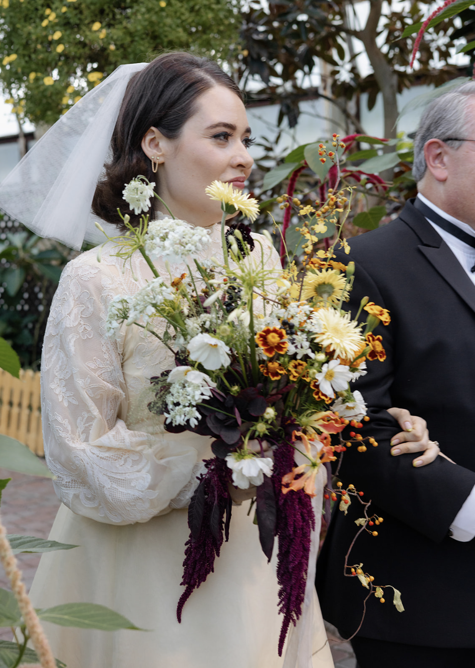 A bride with dark brown hair wearing a lace wedding dress, holding a colorful bouquet of flowers including yellow, orange, white, and purple blooms, standing outdoors with trees and greenery in the background.