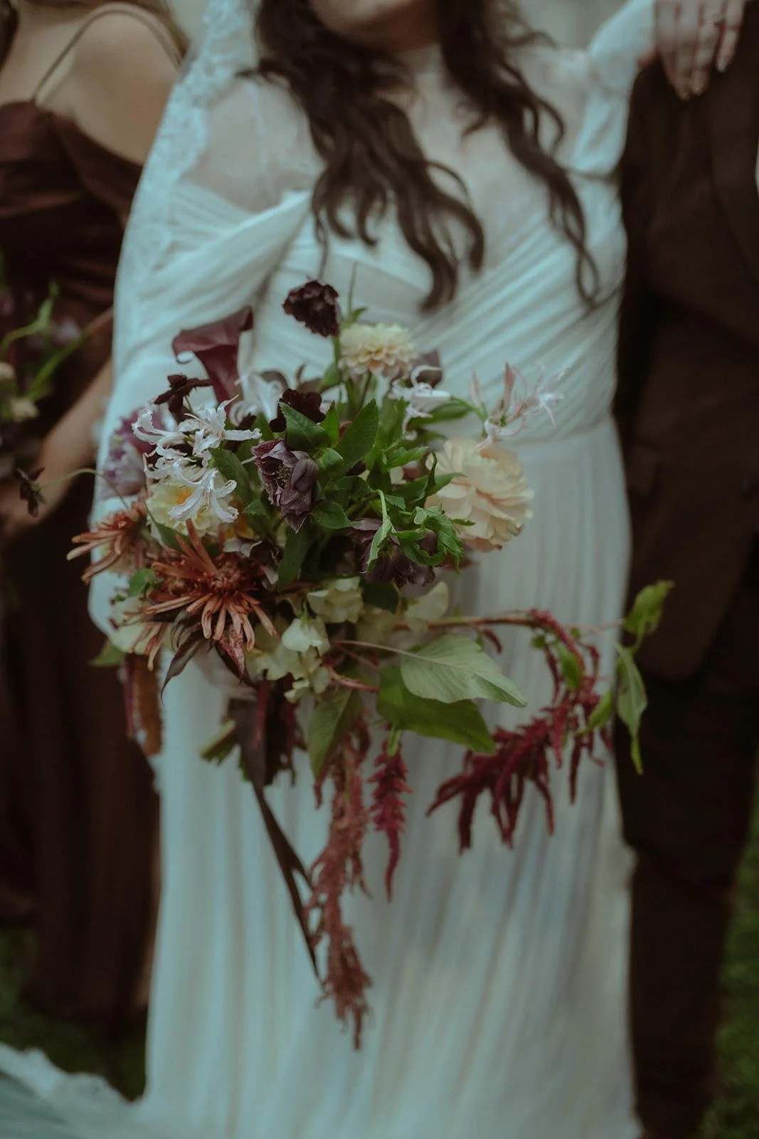 A woman in a white dress holds a large bouquet of mixed flowers including purple, white, and pink blossoms with green leaves and trailing red foliage.