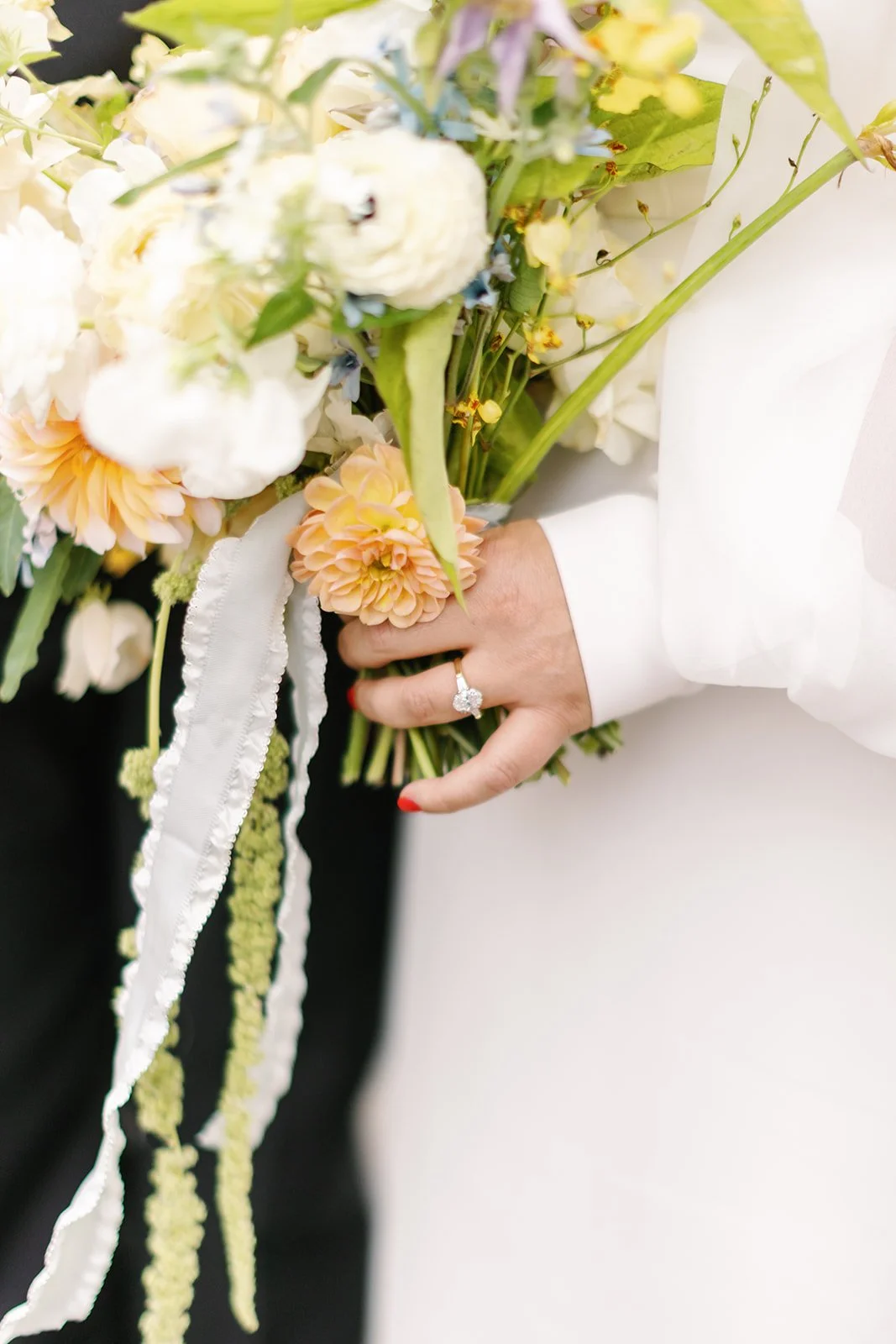 Person holding a bouquet of various flowers, with a focus on a ring on their finger, wearing a white long-sleeve garment.