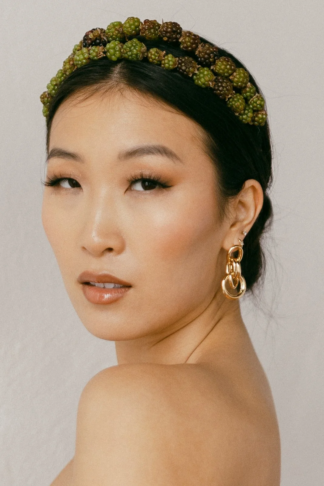 Close-up of a woman with smooth skin, wearing bold eye makeup and glossy lip color, accessorized with large gold hoop earrings and a headband decorated with black and green berries.
