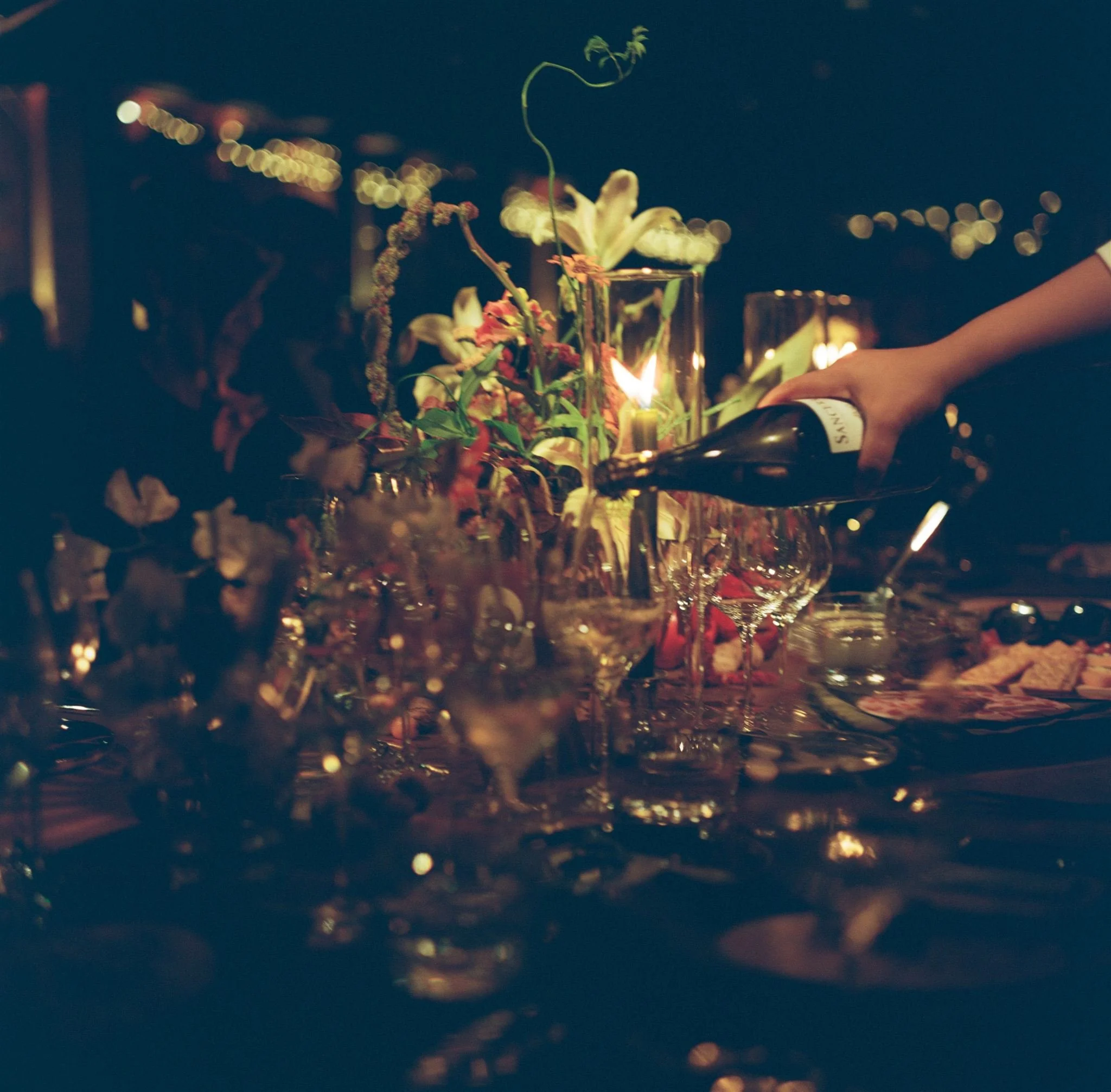Person pouring wine at a dinner table decorated with flowers and candlelight during evening or nighttime.