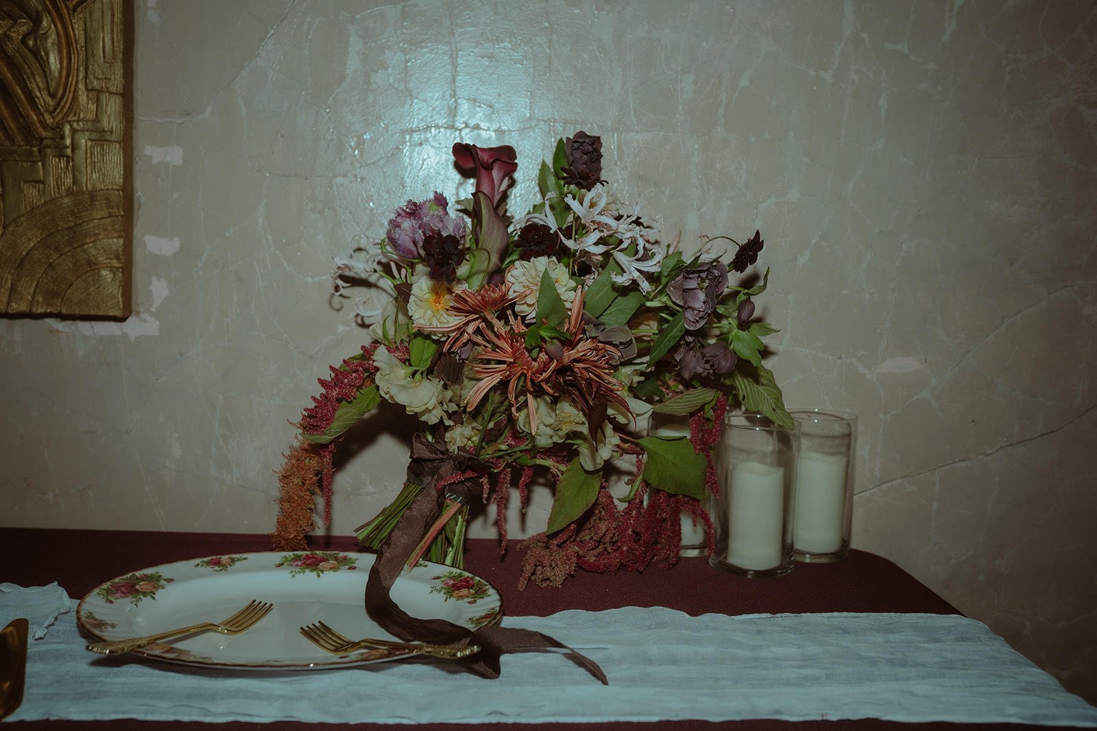 A floral centerpiece with pink, white, and purple flowers on a table, flanked by three white candles in glass holders, with a decorative plate and gold forks in the foreground, and a textured wall in the background.