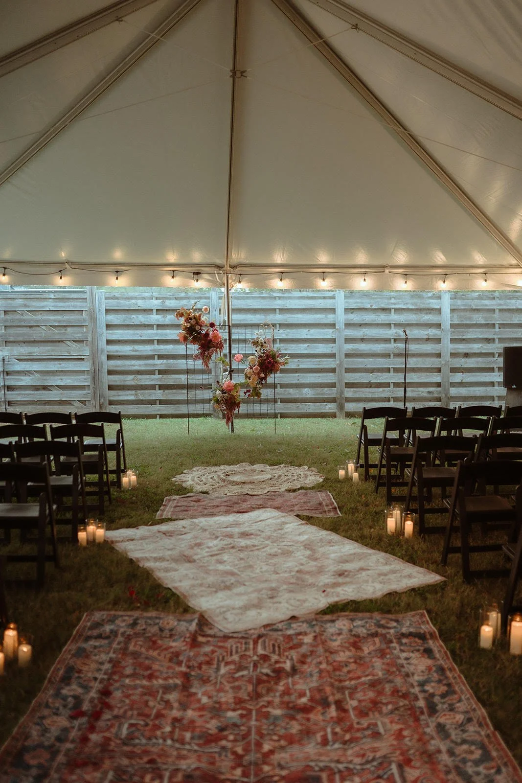 A decorated outdoor wedding ceremony setup inside a tent with string lights, floral centerpieces, a carpeted aisle with candles, and black chairs on either side.